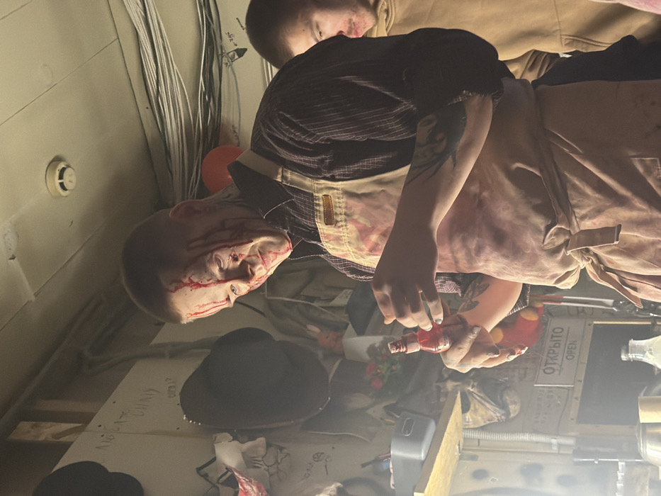 The image displays an indoor scene, likely a themed event or a staged setup, featuring two individuals amidst a cluttered environment. The image is oriented sideways, but will be described as if upright.
In the foreground, a person is prominently featured, their face covered in what appears to be fake blood, streaming down from their forehead and around their eyes and nose. They have a short haircut and a pale complexion. This individual is wearing a dark, striped short-sleeved shirt, over which a light brown or tan apron is worn. The apron also shows splatters or stains resembling blood. Their left arm, visible, appears to have a tattoo. They are holding a small, red plastic bottle in their left hand, which might contain more fake blood or be a prop. Their expression is difficult to discern due to the blood, but their gaze is directed slightly upwards and to the right.
Behind and slightly to the left of the foreground person, another individual is partially visible. They are wearing a light brown or tan hooded sweatshirt or jacket. Only their head and upper torso are seen, and their face is mostly obscured.
The setting appears to be a room with light-colored walls. Along one wall, numerous white and black electrical wires or cables are neatly bundled and run vertically. To the left of the foreground person's head, a white smoke detector or similar device is mounted on the wall. Further back in the scene, several objects are visible on what appears to be a table or surface. These include a black wide-brimmed hat (possibly a cowboy hat style), some crumpled fabric or paper, and what looks like a small vase with red artificial flowers. A red balloon is also partially visible floating near the ceiling.
Further back, on another surface or counter, various bottles and containers are scattered, including a clear bottle and a red and yellow squeeze bottle. A small wooden block or item is also seen.
A sign with text is clearly visible in the mid-ground. The text reads "ОТКРЫТО" in Cyrillic script, with "OPEN" written below it in English. Below this sign, there's a glimpse of a wall with what appears to be faint graffiti or handwritten text, though it's largely unreadable due to the lighting and angle. One segment might contain the word "NIGHT".
The overall atmosphere suggests a horror-themed or theatrical event, possibly an escape room or a Halloween attraction, given the fake blood and props. The lighting is somewhat dim, contributing to the moody ambiance.