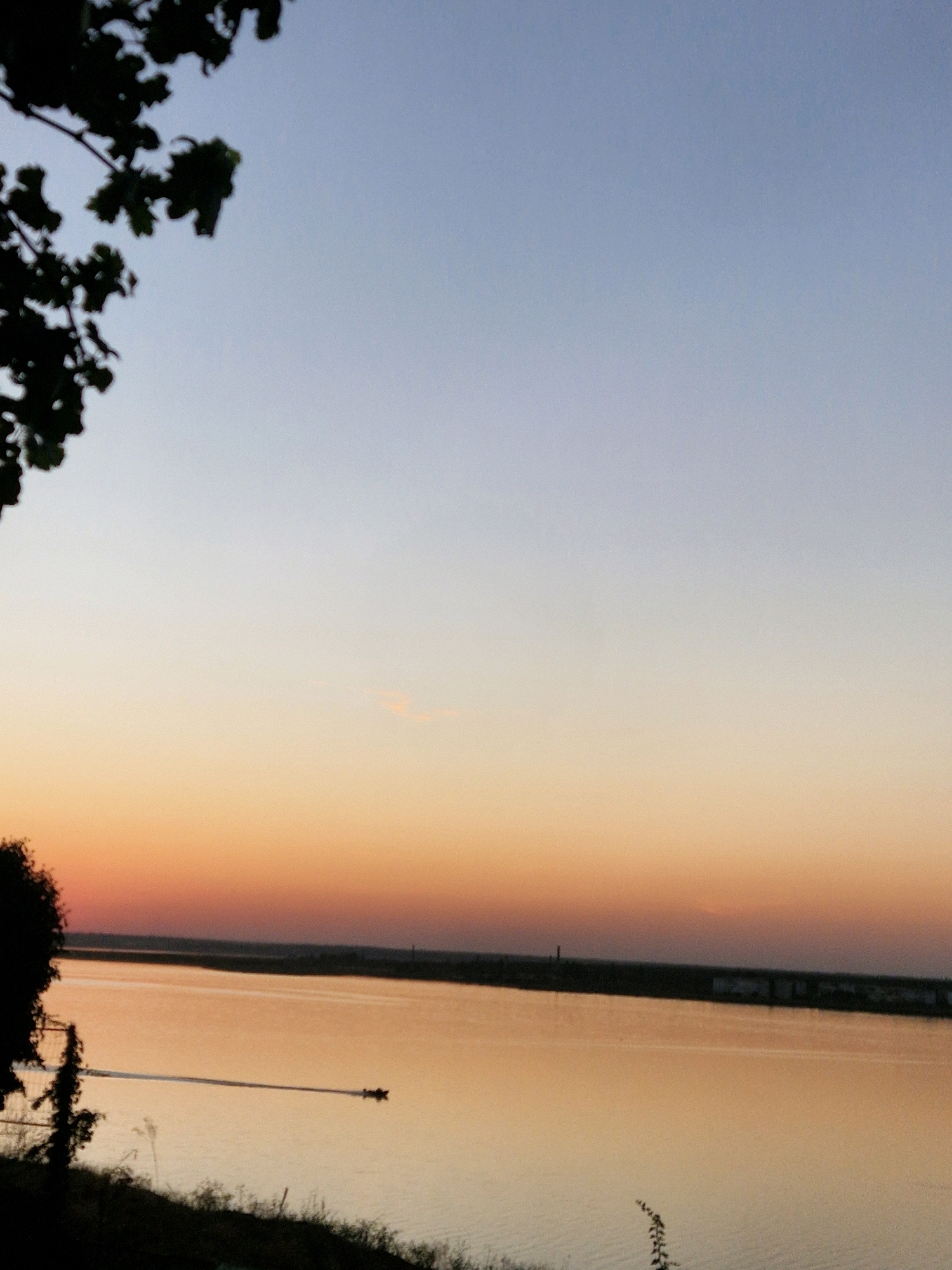A wide shot captures a tranquil scene by a river at sunset. The sky transitions from a pale blue at the top to a soft orange and pink hue near the horizon. Silhouetted foliage from a tree frames the upper left corner of the image, and the dark outline of a distant shoreline runs horizontally across the middle ground. On the water, a small boat is visible, leaving a subtle wake as it moves from left to right. The river's surface reflects the warm colors of the sunset, creating a serene and peaceful atmosphere. This image was taken in the city of Mykolaiv, Ukraine.
