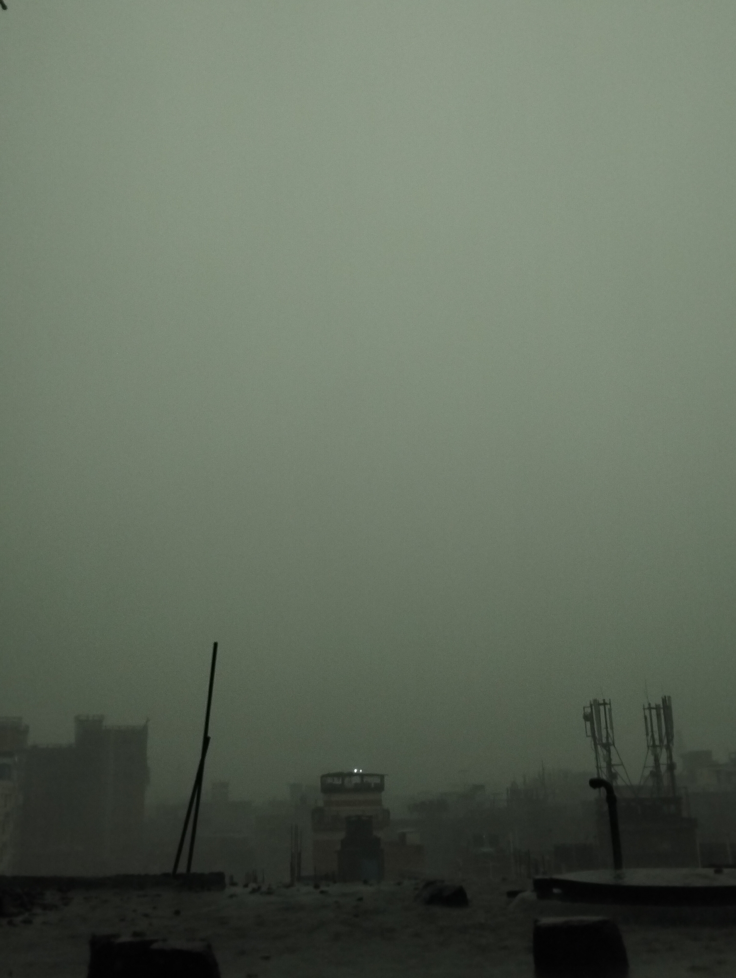 The image shows a cityscape in Dhaka, Bangladesh, heavily obscured by smog or fog. Dominating the view are buildings and structures fading into the haze. In the foreground, there is a rooftop with scattered debris and protruding black bars or poles. Further back, indistinct buildings and communication towers are visible. The overall ambiance is muted, with a lack of distinct colors and a pervasive gray-green tint due to the atmospheric conditions.