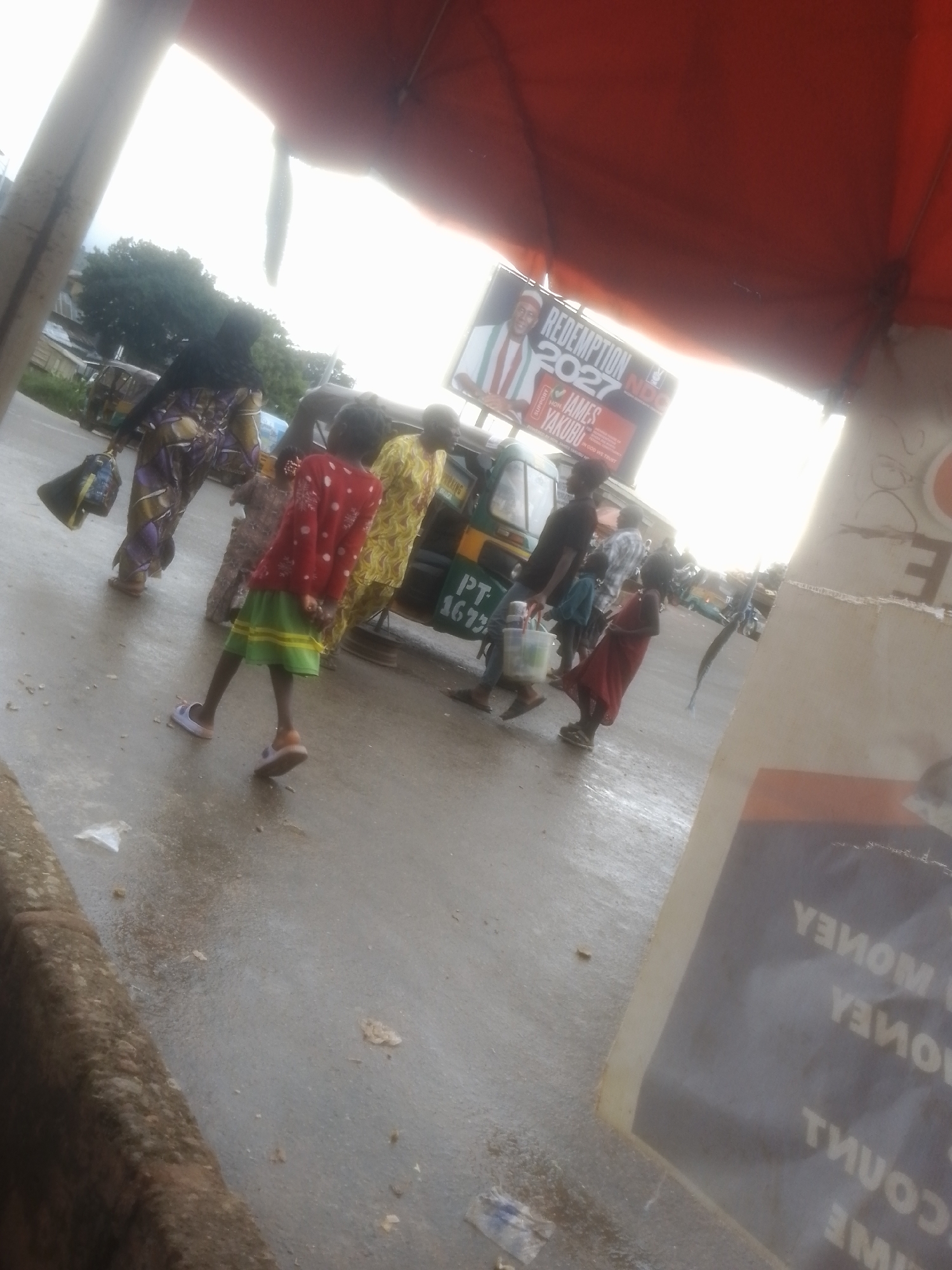 In an outdoor scene in Gwong, Nigeria, a group of people are gathered near a red awning covering. A young girl in a red shirt with white polka dots and a green skirt stands on the wet asphalt. To her left, an adult wearing a colorful patterned outfit carries a bag. Behind them, a three-wheeled vehicle, possibly a tricycle or auto-rickshaw, is visible, with a sign on its side that reads "PT 4673". A billboard advertising "REDEMPTION 2027" featuring a man's face is partially visible behind the awning. Other individuals are seen in the background, and some appear to be walking or waiting. The ground is wet, suggesting recent rain. A poster on the right side of the image displays reversed text, with words like "MONEY" and "TIME" discernible.