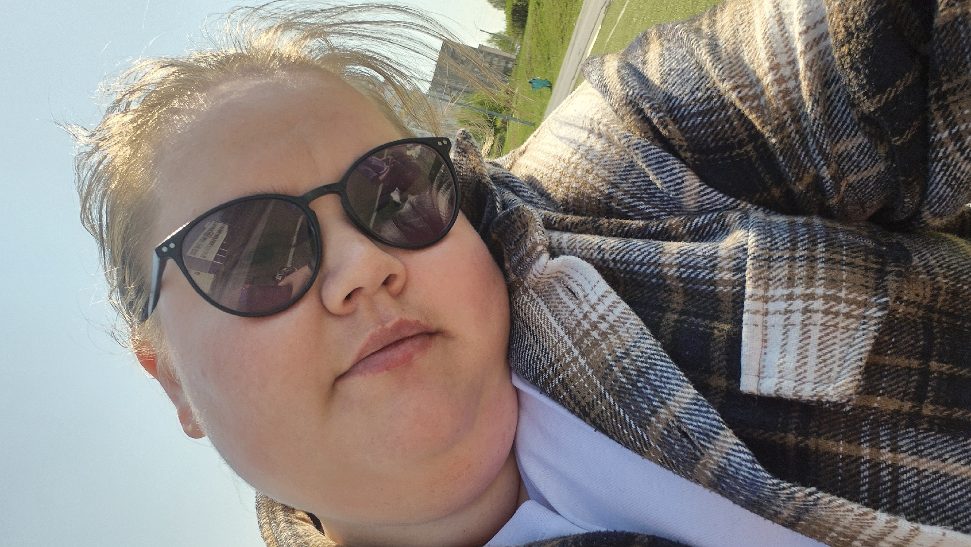 A selfie of a woman wearing black sunglasses and a brown and white plaid jacket. The reflection in the sunglasses shows buildings and possibly a sign. The background appears to be outdoors with a green grassy area and a building in the distance. The photo was taken in Chaikovsky, Russia.