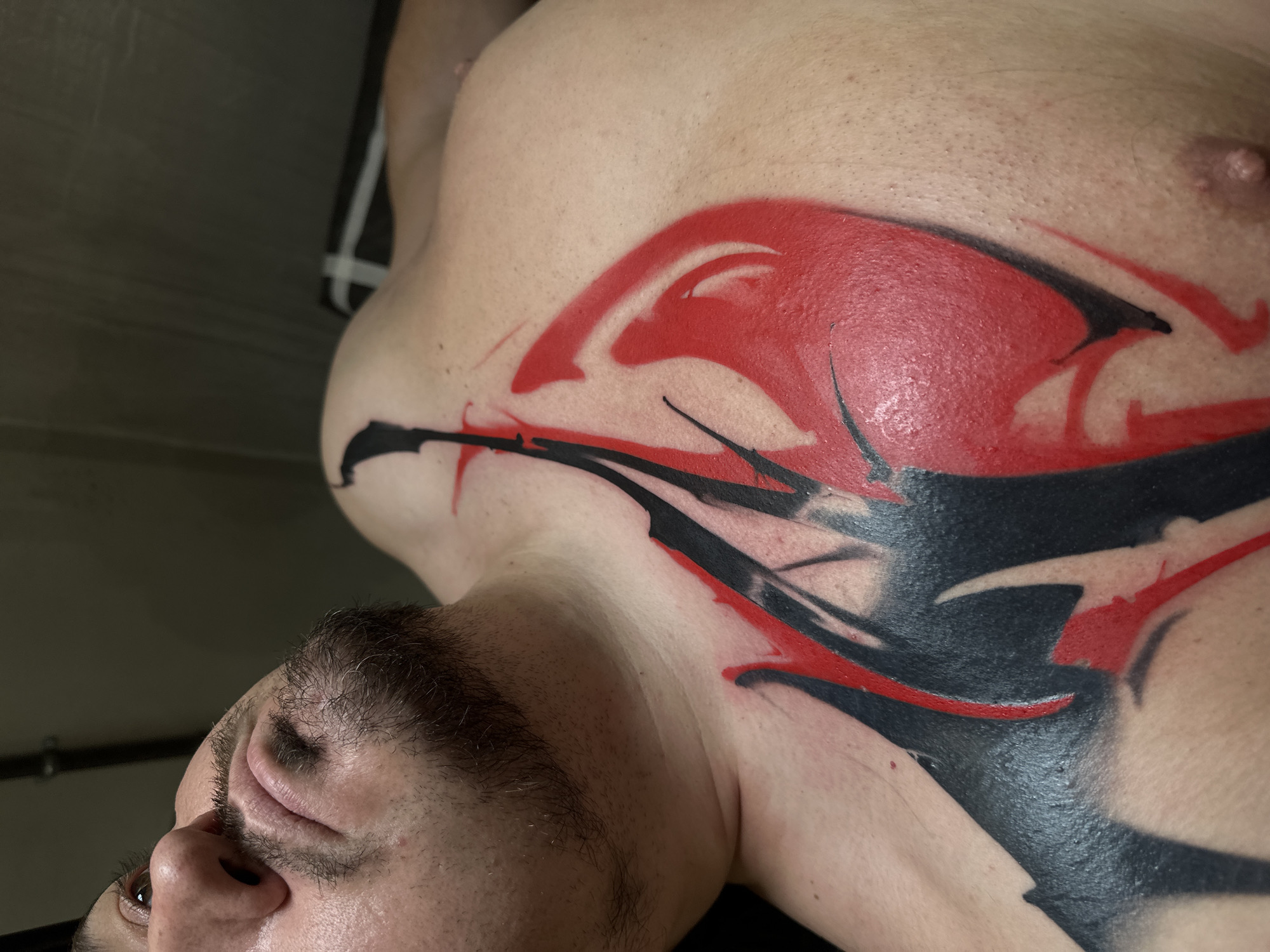 A close-up, slightly angled shot shows a man's upper torso and part of his face. The man has facial hair and is lying down, appearing to look upward. A large, predominantly red and black tattoo covers a significant portion of his chest and extends towards his shoulder. The tattoo has an abstract design with bold strokes and fills. The background is indistinct, but reveals a glimpse of gray wall and a black and white striped item.
There is no information to suggest the location is Moscow, Russia.