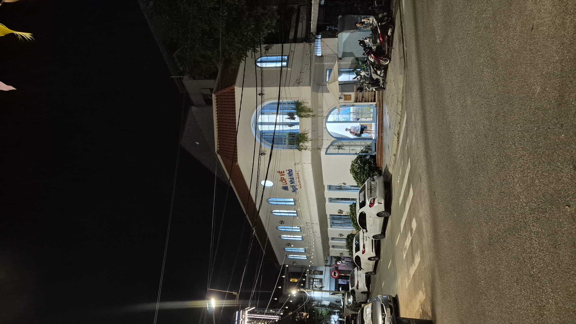 The image captures a night scene on a street in Đà Nẵng, Vietnam. A white, two-story building with a red tiled roof dominates the center of the frame. The building features arched windows, some of which are illuminated, and a prominent sign in Vietnamese that reads "Lớp Vẽ - Ngôi Nhà Nhỏ" (Art Class - Small House). The ground floor has a large open entrance with double doors, and a person is visible standing inside.
In front of the building, several white sedans are parked along the street. To the right, a collection of motorcycles and scooters are parked. The street itself is paved, and a pedestrian crossing is marked with white stripes. Overhead, power lines and streetlights are visible against the dark night sky. The surrounding environment suggests an urban setting with other buildings and trees present, though they are less distinct in the nighttime lighting. The overall atmosphere is calm and serene, typical of a quiet street in the evening.