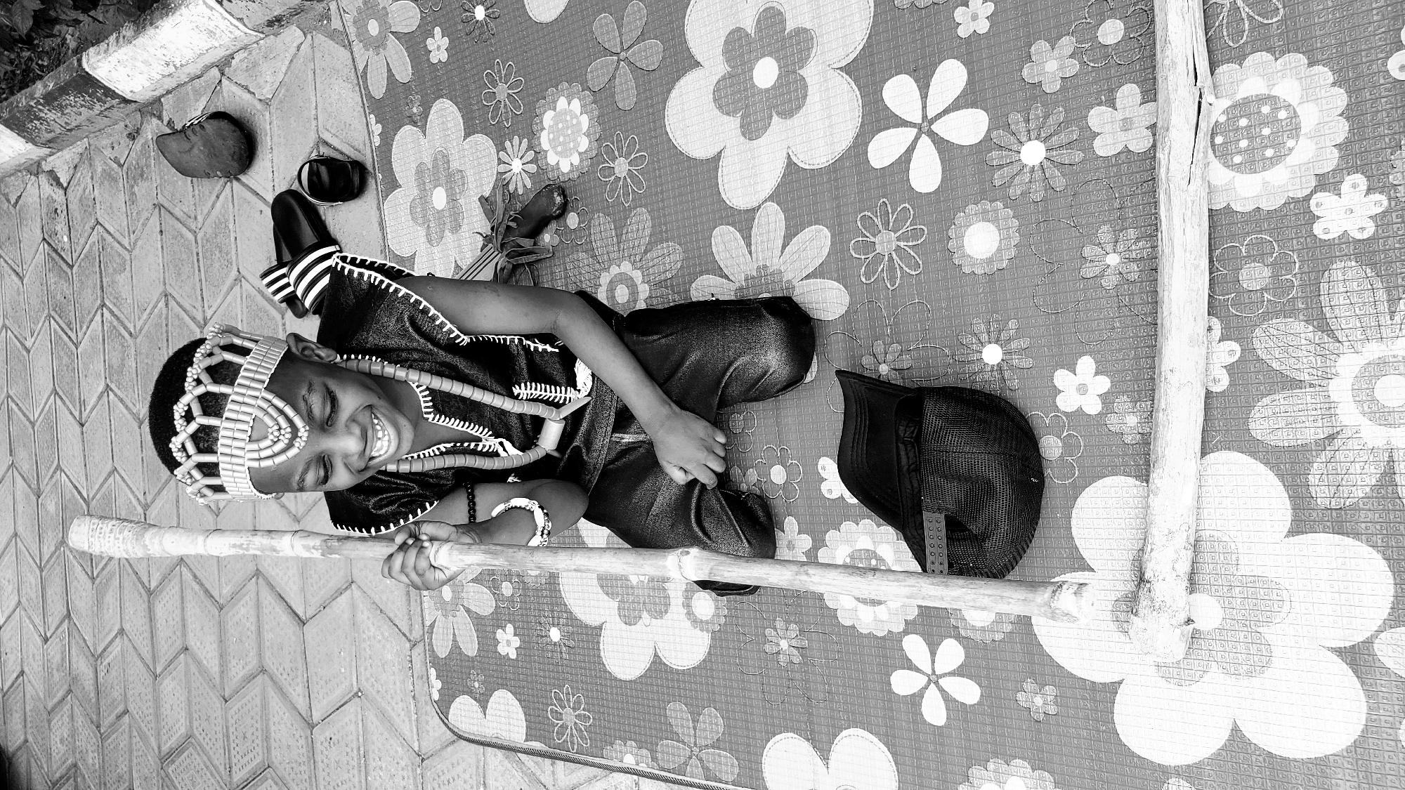 A child in traditional attire lies on a floral patterned mat in what appears to be Sabon Tasha, Nigeria. The child is smiling and holding a bamboo stick. He or she is wearing a beaded headpiece, a beaded necklace, and a traditional garment with beaded accents. To the side are shoes and a baseball cap. The mat is on a tiled surface next to a painted curb.