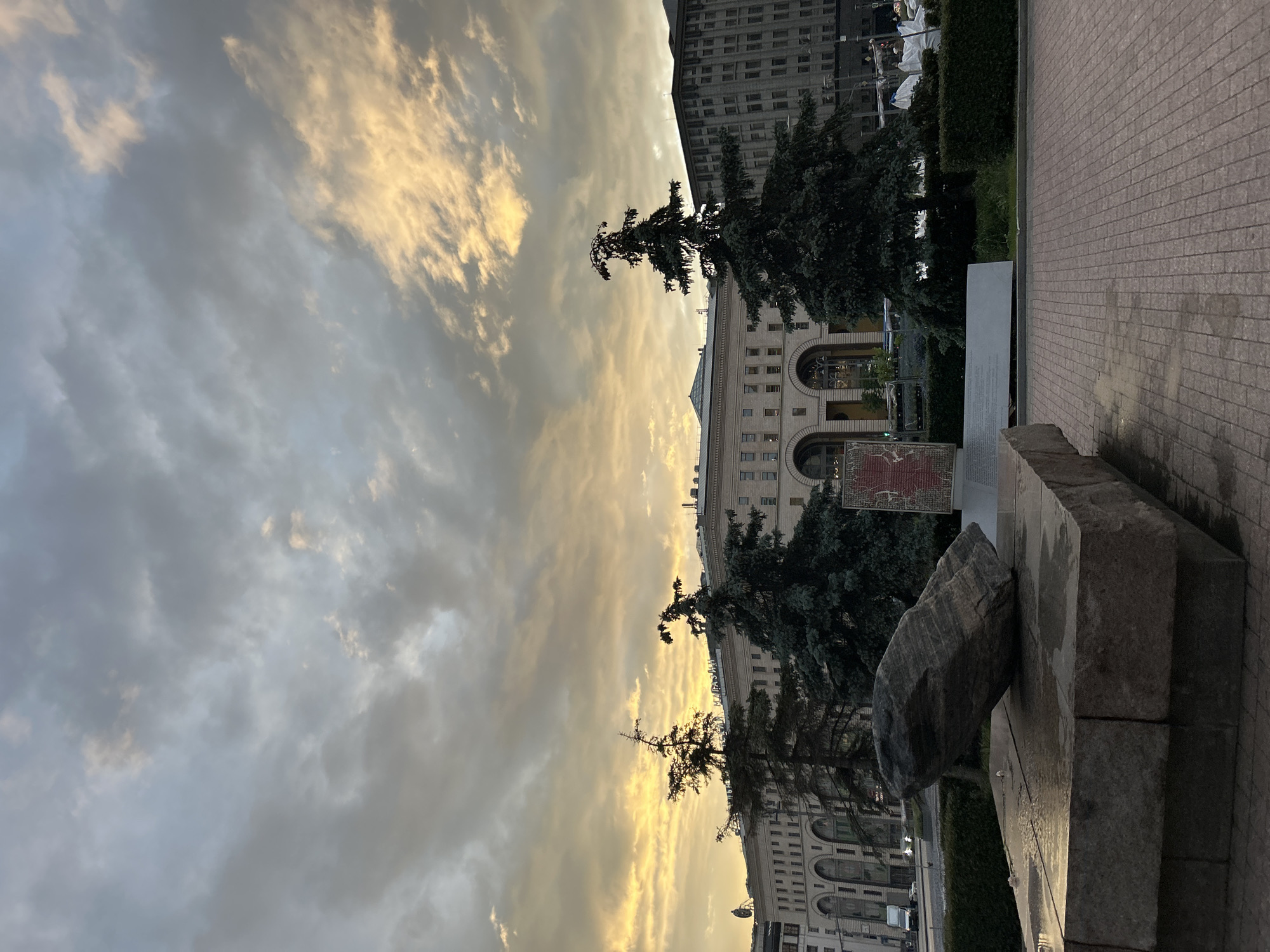 The image depicts a cityscape at dusk. The sky is filled with gray and yellow clouds. In the midground, there are two evergreen trees framing a light-colored building with arched windows. To the right is a stone structure, appearing to be part of a memorial. The ground is paved with rectangular stones. Another building is visible in the background. The location is in the city of Unknown, Unknown.