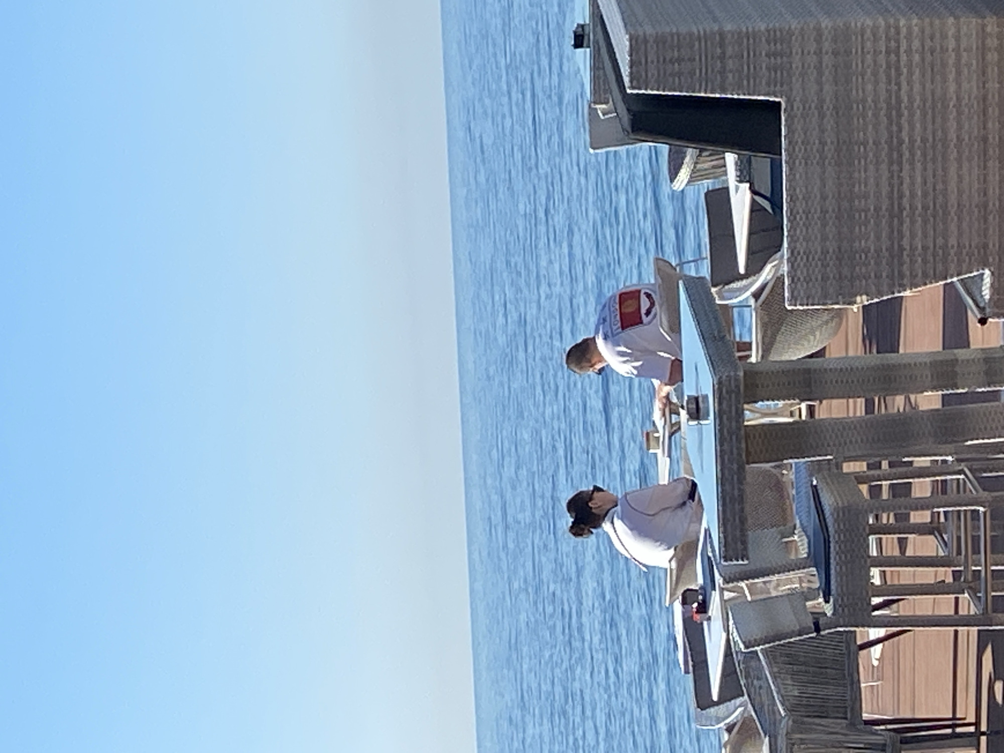 Two people are on a deck with wicker furniture, possibly a cafe or restaurant, overlooking the sea under a clear blue sky. The location is likely Budva, Montenegro, given the presence of the Adriatic Sea. One person, with dark hair pulled back, is wearing a light-colored shirt. The second person is wearing a white shirt with a logo or design on the back.