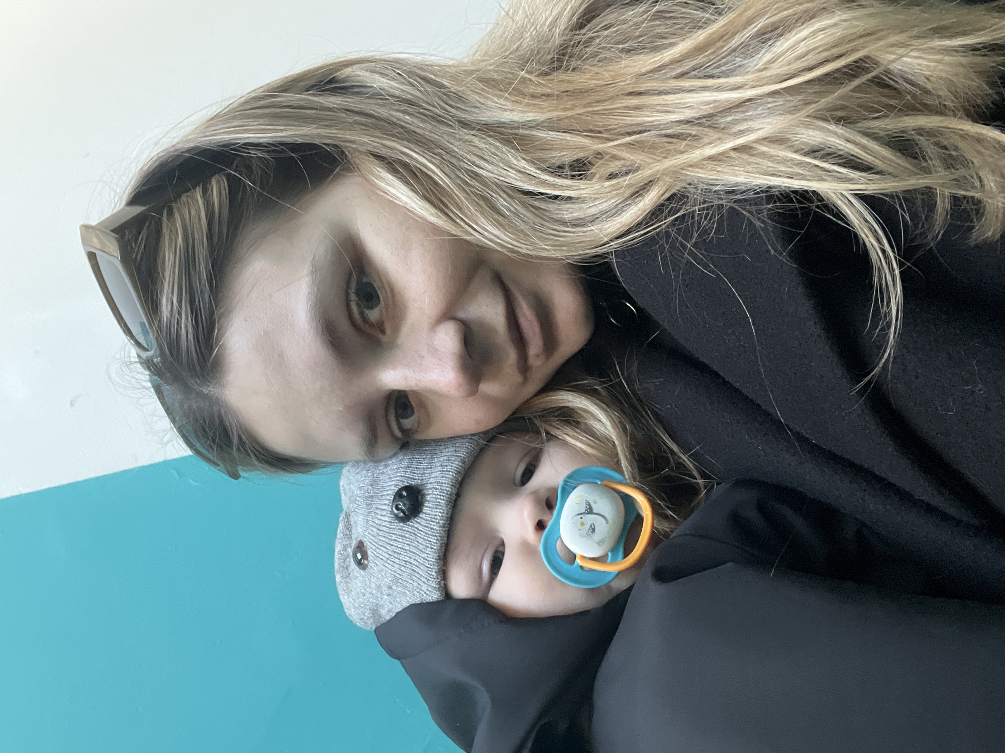 An adult female and an infant are captured in a close-up indoor portrait. The female, positioned on the right, has long, blonde hair and is wearing a dark garment. Sunglasses are resting on top of her head. She looks directly at the camera. The infant, held by the female on the left, wears a grey knitted hat with two dark spherical elements on top, resembling animal ears, and a black hooded coat. A blue and orange pacifier, featuring a white circular shield with a cartoon design, is in the infant's mouth. The infant's gaze is directed slightly towards the camera. The background consists of a teal-blue wall or surface on the left and an off-white wall or surface on the right, indicating an interior setting. The specific location within the city of Chaikovsky, Russia, is not discernible from the visual information provided.