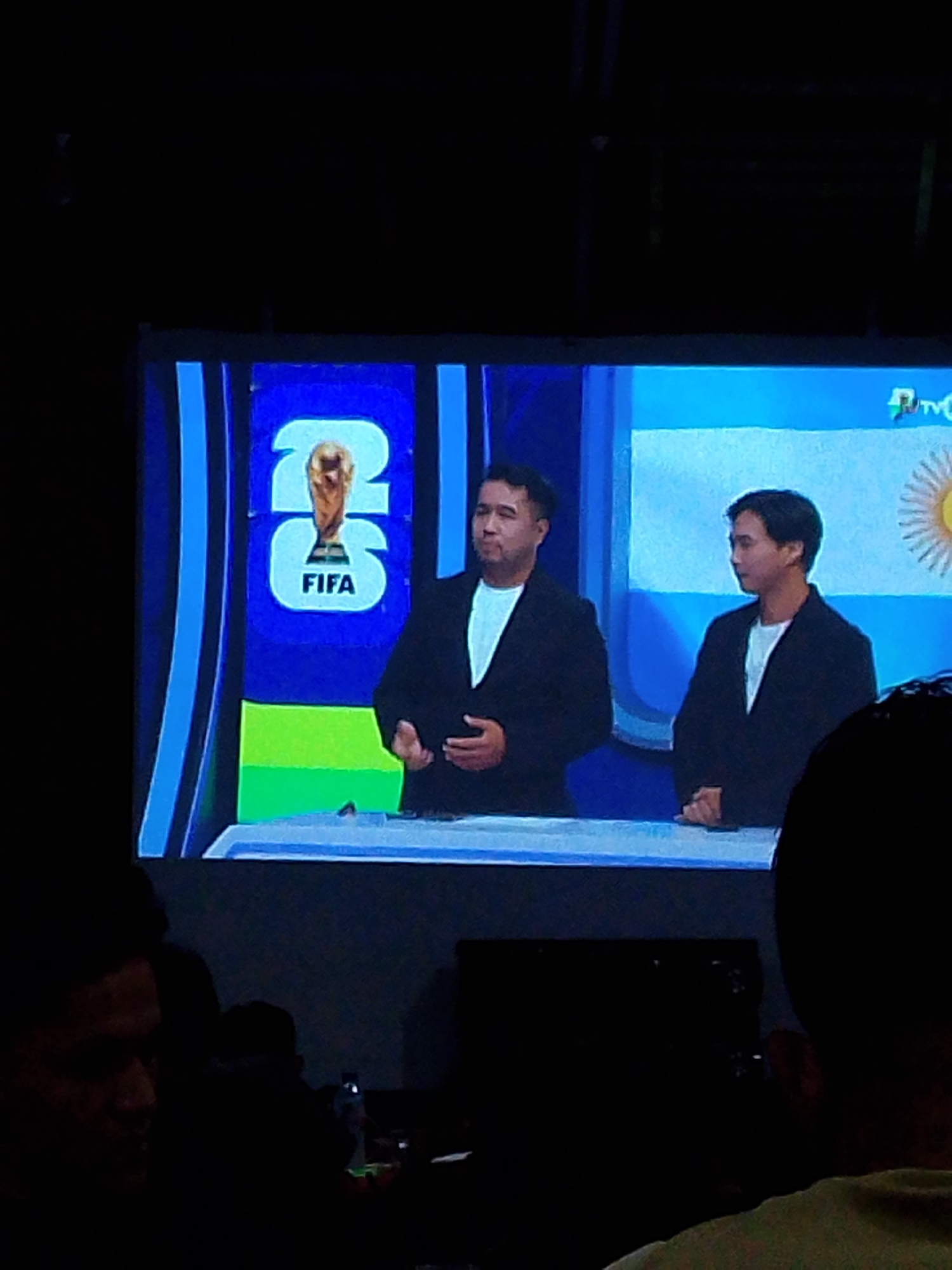 The image shows a large screen displaying a broadcast. On the screen, two men in black blazers over white shirts are visible. The man on the left is slightly older with a fuller face, and the man on the right is younger with a more slender face. Behind them, to the left, is a graphic featuring a trophy and the text "26 FIFA". To the right, a portion of the Argentinian flag is visible. The screen appears to be in a dark environment, possibly a room with an audience, as suggested by the dark silhouettes in the foreground. The broadcast is likely related to a sporting event, possibly the FIFA World Cup. The location is identified as Meunasahmee, Indonesia.