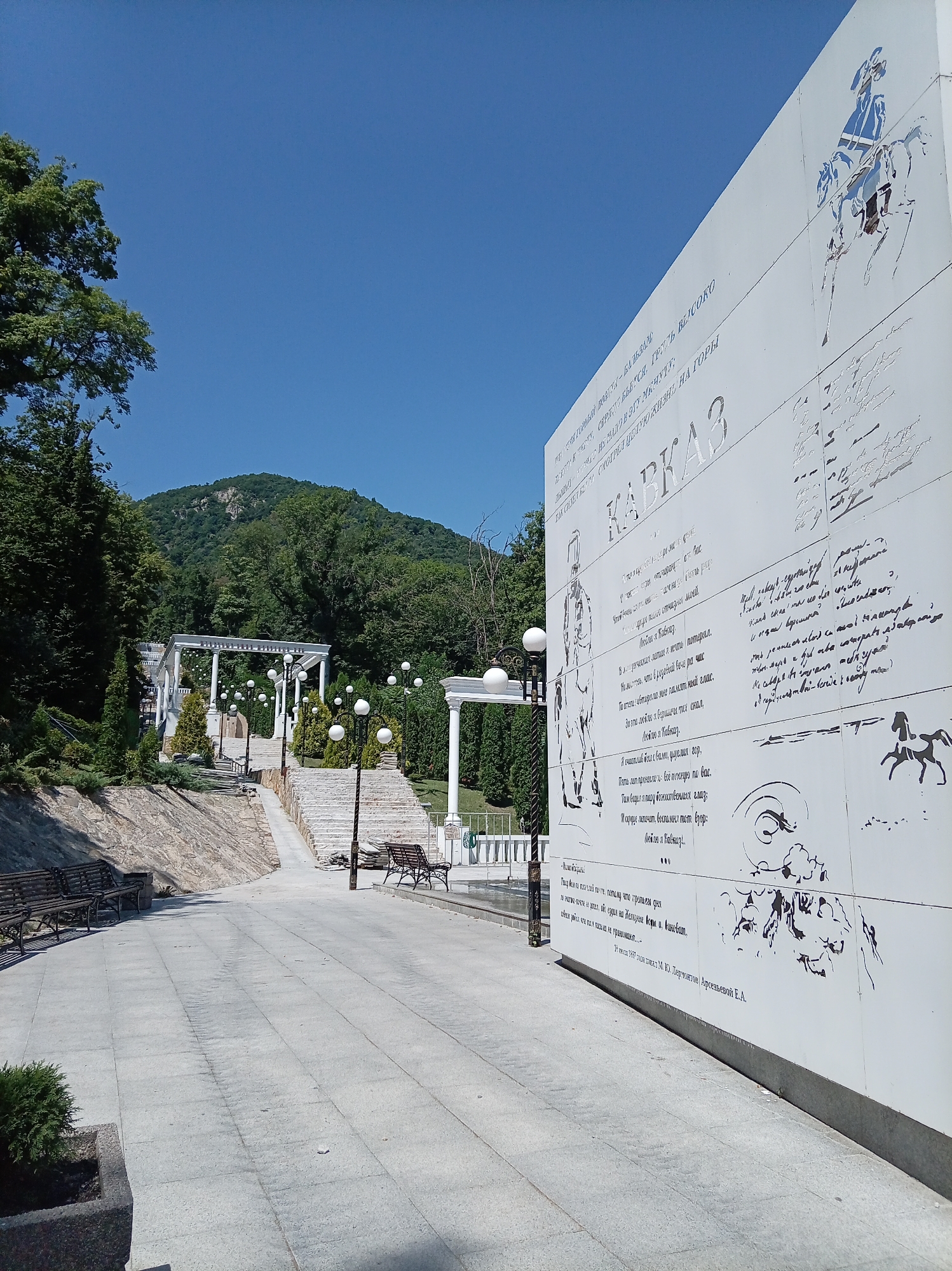 The image shows a view of the monument "Taman Divizion" in the city of Zheleznovodsk, Russia. The monument is a large white wall with bas-reliefs and inscriptions. The wall is decorated with a drawing of a man on horseback and several lines of text. In the background, there is a lush green mountain and a clear blue sky. The foreground shows a paved walkway lined with benches and lampposts. The overall scene is peaceful and serene, with a strong sense of history and culture.