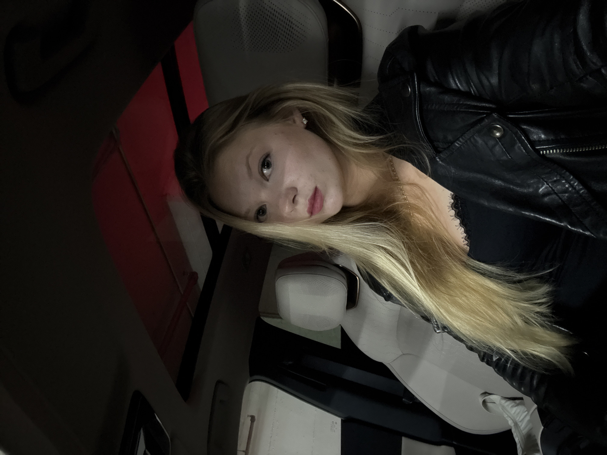 A young woman with blonde hair is reclining in the passenger seat of a car. She is wearing a black leather jacket and a black top. The interior of the car is light-colored with a sunroof visible. The car appears to be stationary. The location is unknown; there is no indication of Tel Aviv, Israel, or any other specific location.