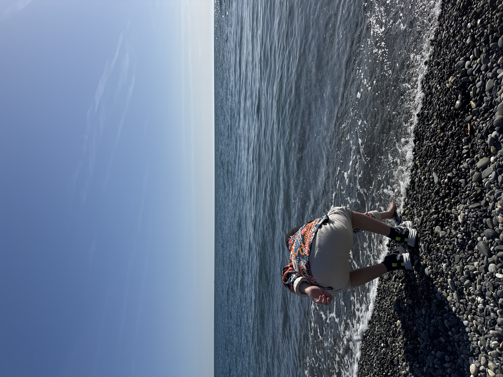 The horizontally oriented image captures a serene daytime scene at a pebbly beach, likely in Sochi, Russia. A person is positioned on the right side of the frame, bent over and interacting with the shallow water at the edge of the shore. They are wearing light-colored shorts or trousers and a vibrantly patterned, short-sleeved shirt featuring red, orange, blue, and green designs. Their bare feet are partially submerged in the foamy water, and their left hand is near the water's surface, suggesting they are wading or reaching for something. A pair of black and white sneakers with distinctive neon yellow accents rests on the dark pebbles next to their right foot.
The beach is composed of numerous dark, smooth, mostly black and grey pebbles, with some appearing wet from the breaking waves. To the left of the beach, the calm, dark blue-grey water of the sea or ocean stretches towards a distant horizon. Gentle white-foamed waves lap at the shore around the person's feet. Above the water, a clear, bright blue sky dominates the upper portion of the frame, with a faint, wispy cloud or contrail visible in the upper left. The bright sunlight indicates it is daytime, casting subtle shadows from the person and the pebbles. No discernible text is visible in the image.