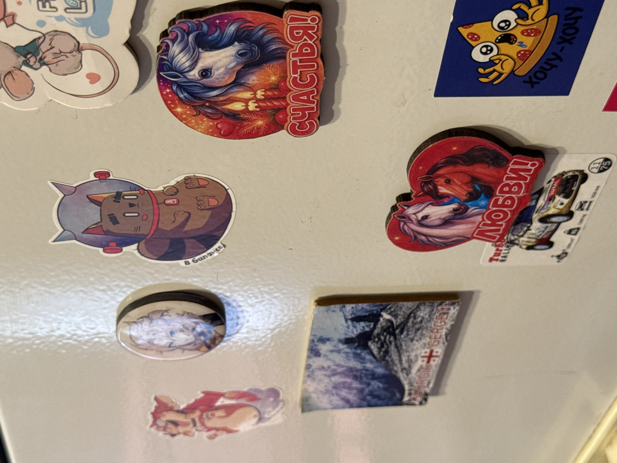 The image shows a close-up of a refrigerator door adorned with various stickers and magnets. The items include cartoonish animal figures, a depiction of horses with text in Russian, a magnet with a landscape scene and text that appears to say "GEORGIA GERGETI," and a sticker of a startled pizza slice with Russian text. The refrigerator door is a neutral, off-white color, and the lighting suggests an indoor environment. There is no discernible context to indicate this is in Яромінскі сельскі Савет, Belarus.