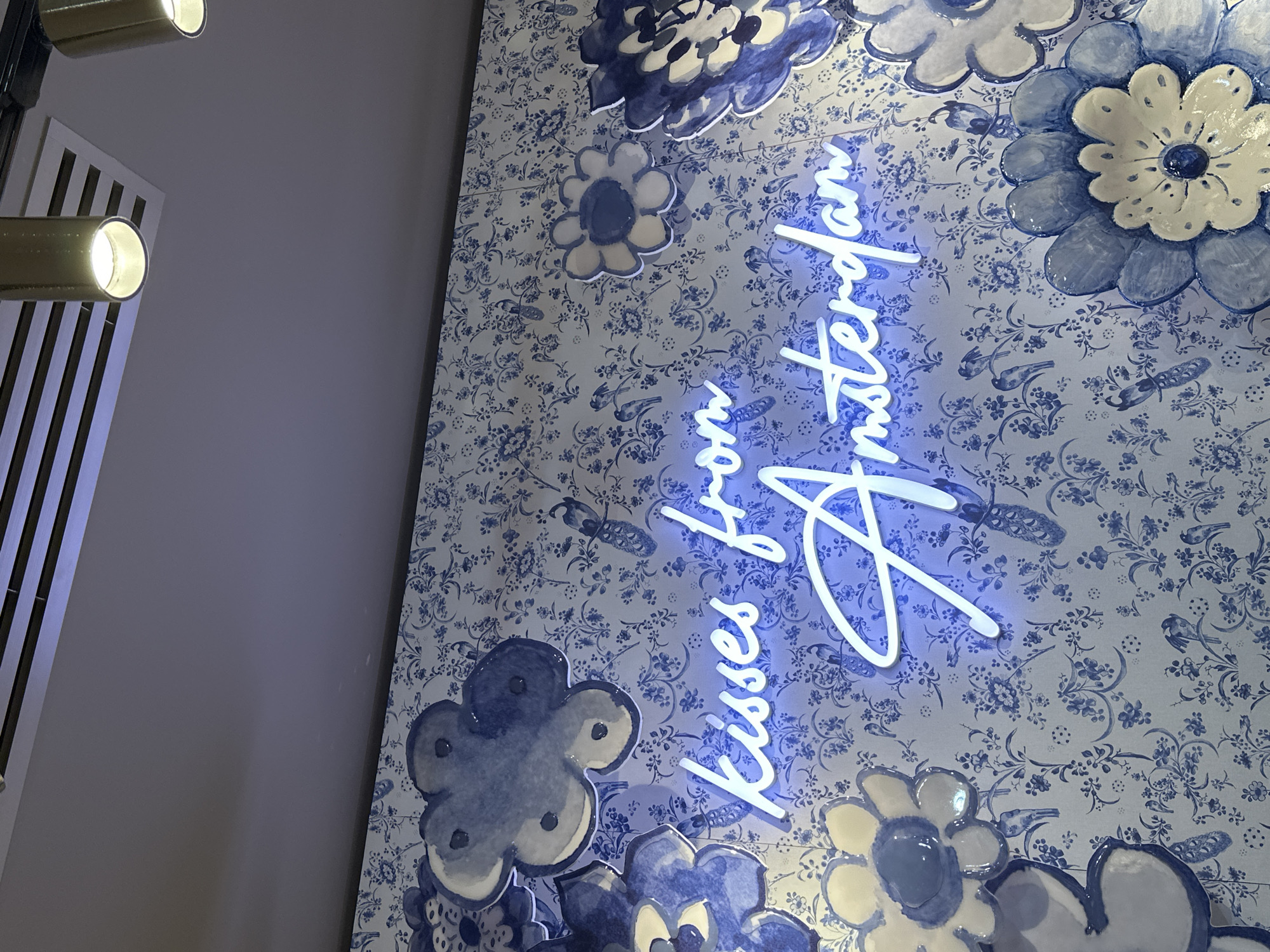 The image shows a wall decorated with blue and white floral wallpaper, reminiscent of Delftware. A neon sign with white light reads "kisses from Amsterdam." Several large, three-dimensional blue and white flowers are attached to the wall. The scene is indoors, with a ceiling visible at the top, featuring lighting fixtures and what appears to be an air vent. The overall aesthetic suggests a decorative element within a public space or establishment in Amsterdam.