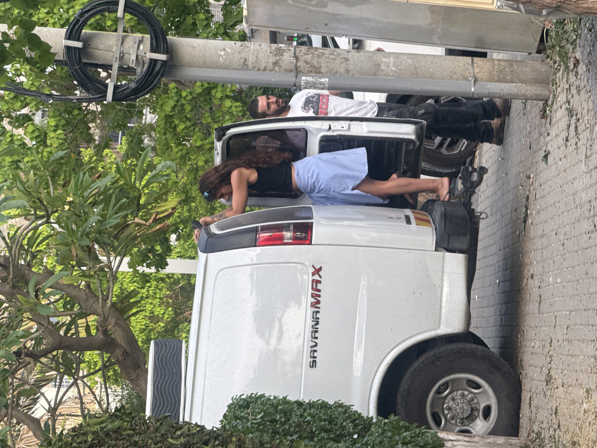 The image captures a scene in Givatayim, Israel, featuring two individuals interacting with a white "SAVANA MAX" van parked beside a brick sidewalk near greenery and a utility pole with coiled black cable. One individual, a woman with long dark hair wearing a black top and light blue skirt, is positioned horizontally across the van's open rear doors, her body supported by her hands and feet. The second individual, a man with a beard wearing a white t-shirt and dark pants, is standing behind her, leaning into the open back of the van as well.