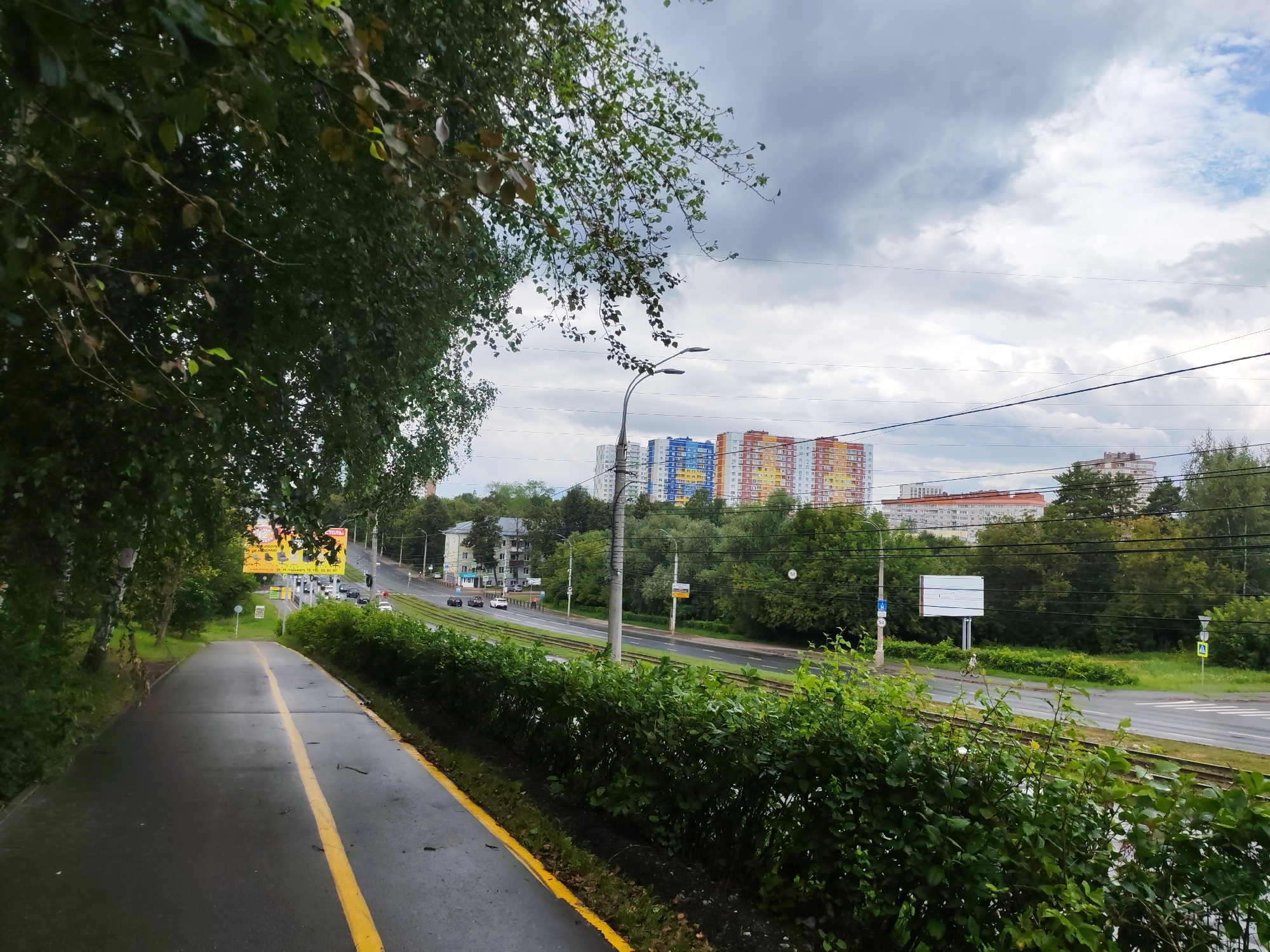 The image depicts a street scene in Izhevsk, Russia, on a cloudy day. In the foreground, a wet asphalt path with a yellow line runs alongside a dense green hedge. Beyond the hedge, a multi-lane road is visible with cars driving on it, along with tram tracks. Further in the background, several multi-story apartment buildings of various colors, including blue, yellow, and orange, are seen behind a line of trees. Power lines are strung across the sky. A street lamp stands on the side of the road.