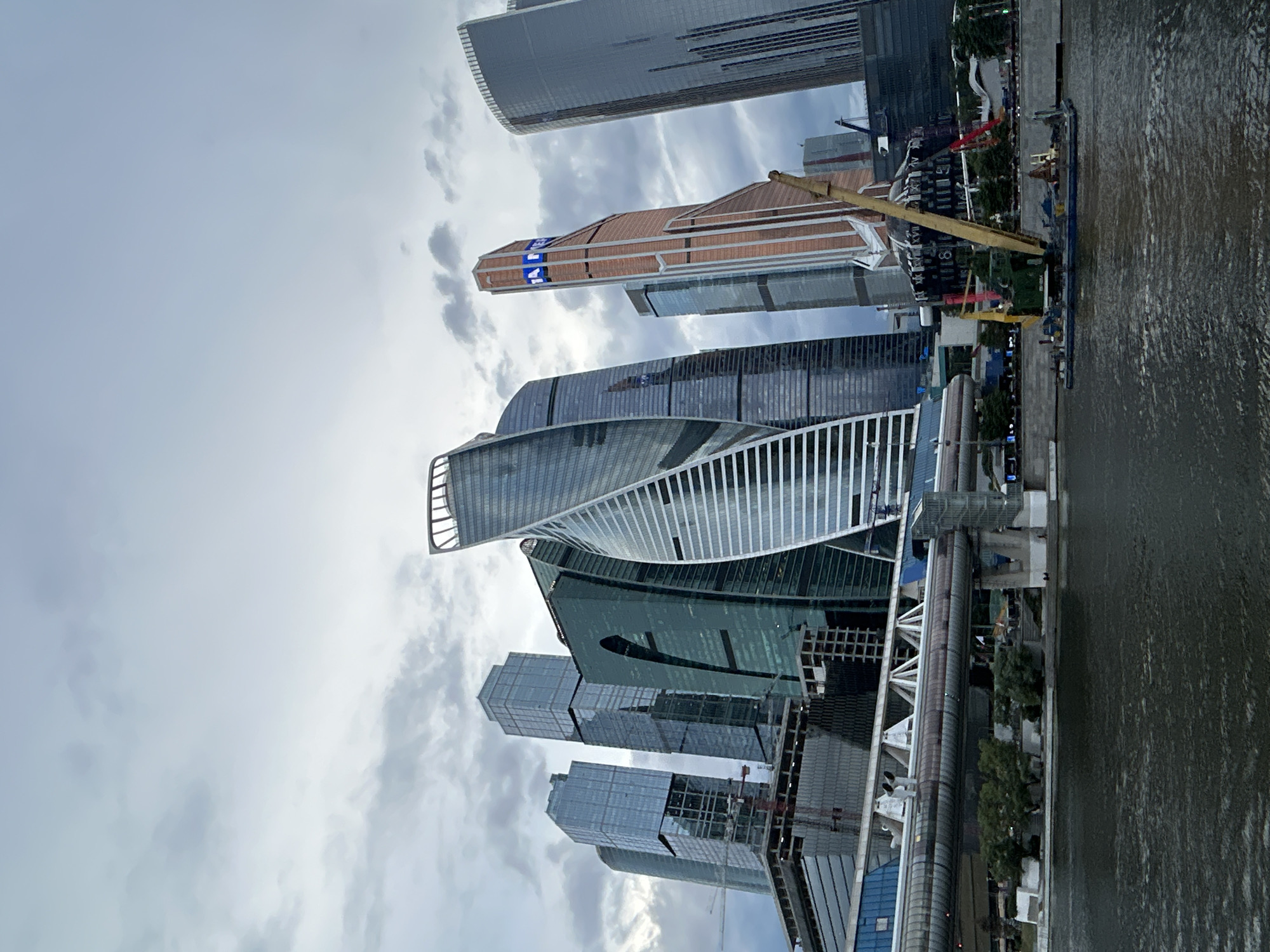 This image captures the Moscow International Business Center, commonly known as Moscow-City, a developing business district in west central Moscow, Russia. The foreground features a dark, choppy river, likely the Moskva River, with a barge carrying a large yellow crane docked along its edge. In the background, a cluster of modern skyscrapers dominates the skyline under a cloudy, overcast sky. The buildings are a mix of glass and steel, with diverse architectural styles, including the distinctive twisting skyscraper of the Neva Towers and the striking copper-colored Federation Tower. A bridge structure with enclosed pedestrian walkways spans across the foreground, connecting different parts of the city. The overall scene depicts a busy urban environment with ongoing construction, indicated by the crane and scaffolding visible on some of the buildings. There are no discernible people or animals in the frame.
