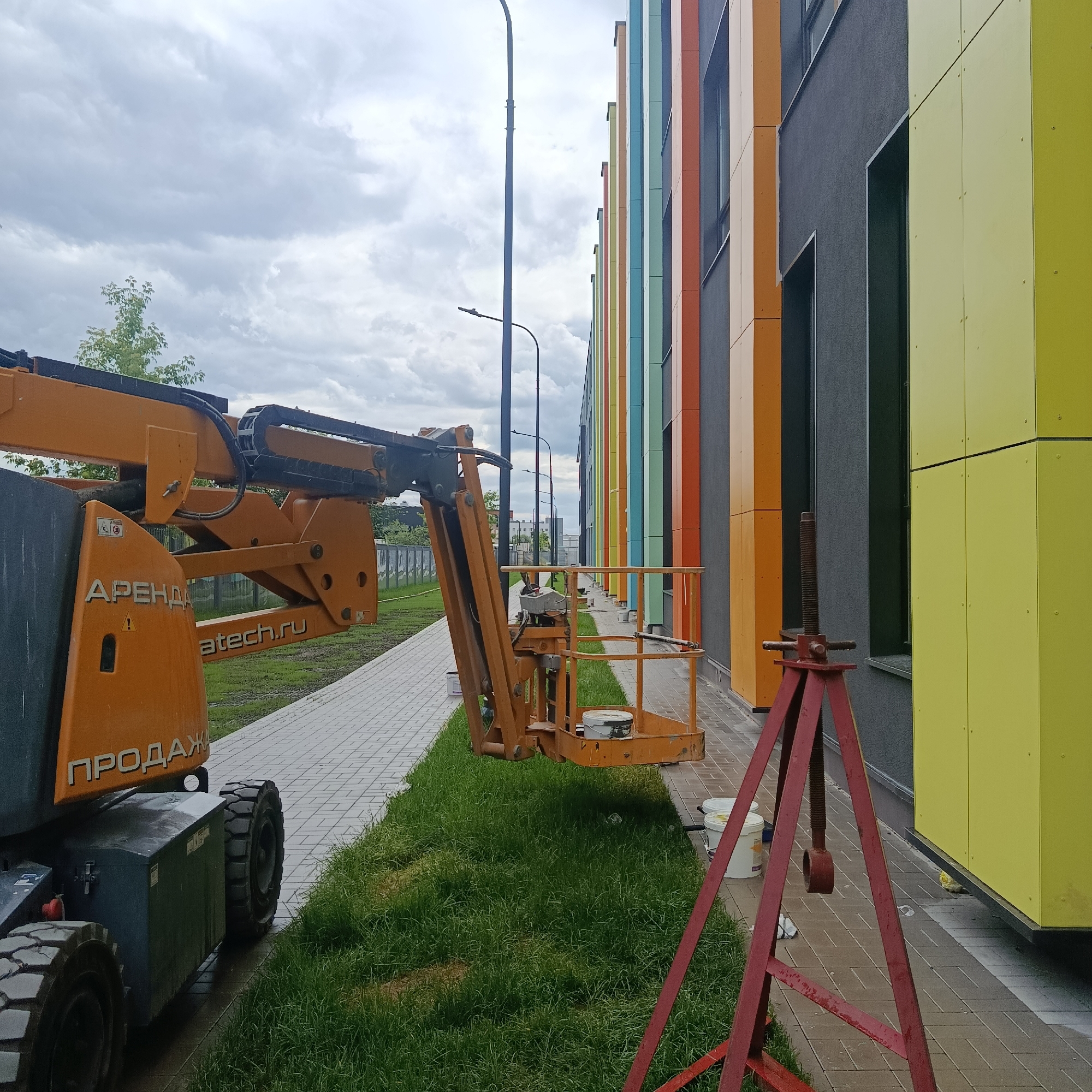 The image depicts an outdoor scene, likely in an urban setting, with an emphasis on construction or maintenance work. In the foreground, a large, orange articulated boom lift is parked on a paved walkway. The lift has visible branding, including "АРЕНДА" (rent) and "atech.ru" on its side. Adjacent to the lift is a red metal tripod stand with a threaded rod, positioned on the grass.
The background features a modern building with a facade composed of vertical panels in various bright colors, including yellow, lime green, teal, orange, and beige, interspersed with dark gray sections. Several windows are visible on the building. A lamppost stands to the left of the building, and a line of similarly styled lampposts recedes into the distance. The ground is covered by a tiled walkway and a strip of green grass. The sky is overcast, with visible clouds. The scene appears to be under construction or undergoing exterior work, suggested by the presence of the boom lift and the tripod stand. There are no people visible in the image. The presence of Russian text on the boom lift suggests the location is likely in Russia. While Moscow is mentioned in the prompt, there are no specific visual cues in the image that definitively place it in Moscow.