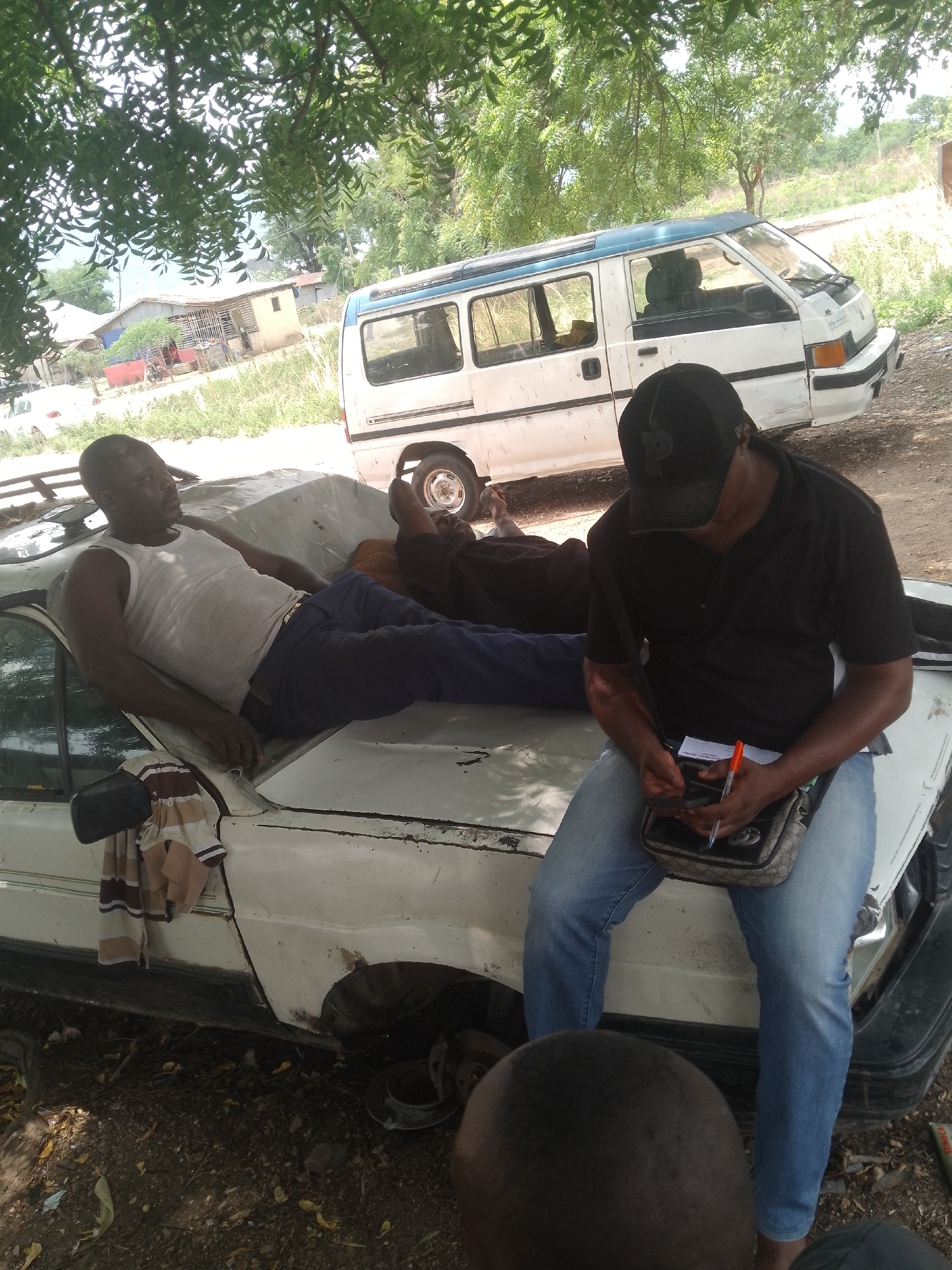The image shows two men in an outdoor setting in the city of Unknown, Unknown. One man is lying on the hood of a white, old car with a white covering, dressed in a white tank top and blue pants. He is reclining with his legs propped up. The second man is sitting on the hood of the car, also wearing blue pants and a black shirt. He has a black baseball cap on and is looking down, writing on a piece of paper with an orange pen while holding a cell phone in the other hand. He has a small crossbody bag on his shoulder. The car appears to be in disrepair, with no tire on one of the wheels. Behind the car, there is a white van with a blue roof. The setting appears to be somewhat rural, with trees providing shade. A building is visible in the background.