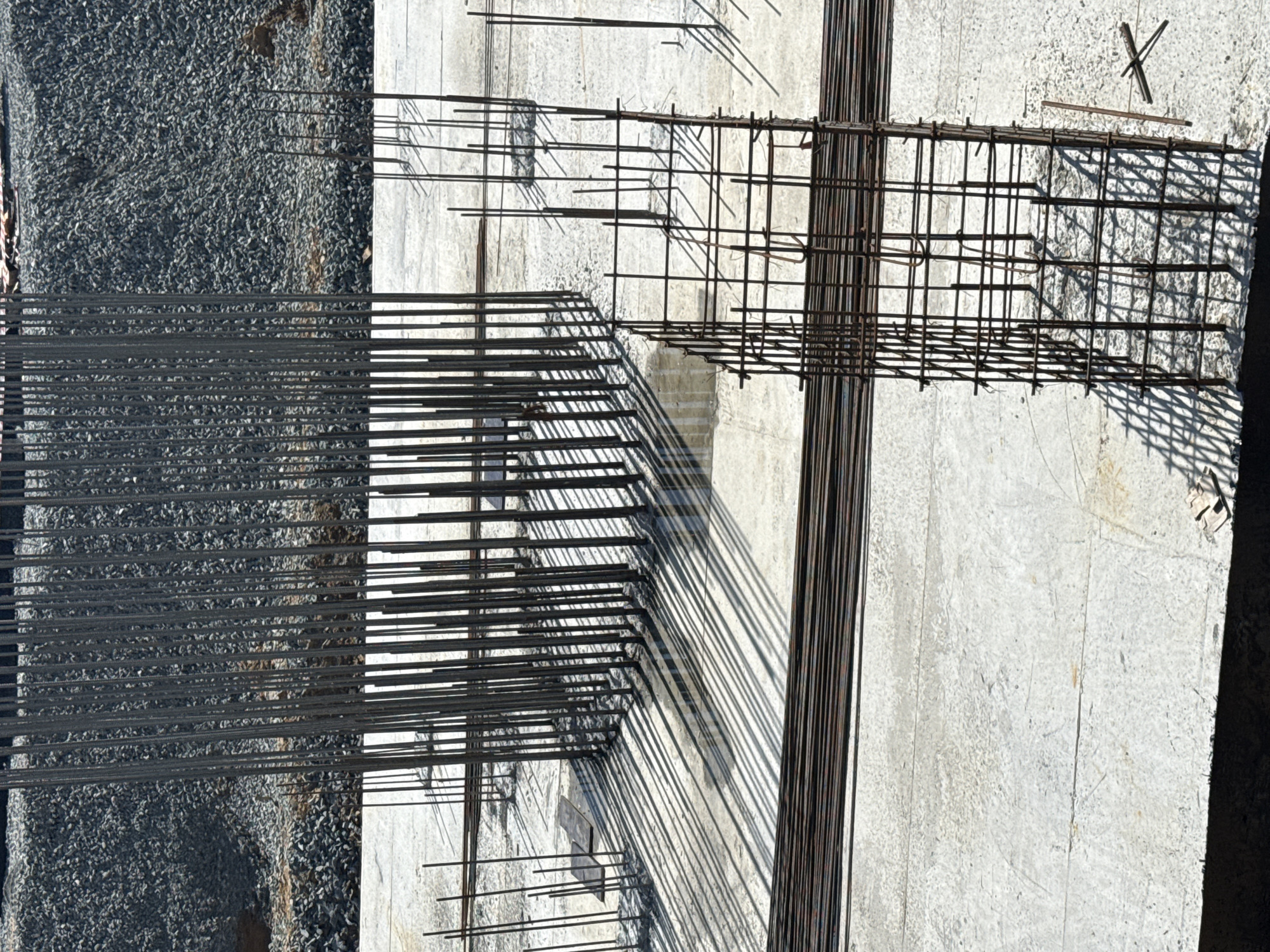 The image depicts a construction site, likely in the city of Leninsk, Russia, featuring the framework for concrete pillars. In the foreground, a square cage of rebar stands on a concrete foundation, with several more rebar cages visible further back. A large, circular cluster of vertical rebar, forming the core of a pillar, dominates the mid-ground. The rebar casts long, distinct shadows across the wet concrete surface, indicating a sunny day. In the background, a large pile of gravel and debris is visible. The overall scene conveys an active construction zone in the early stages of building a structure.