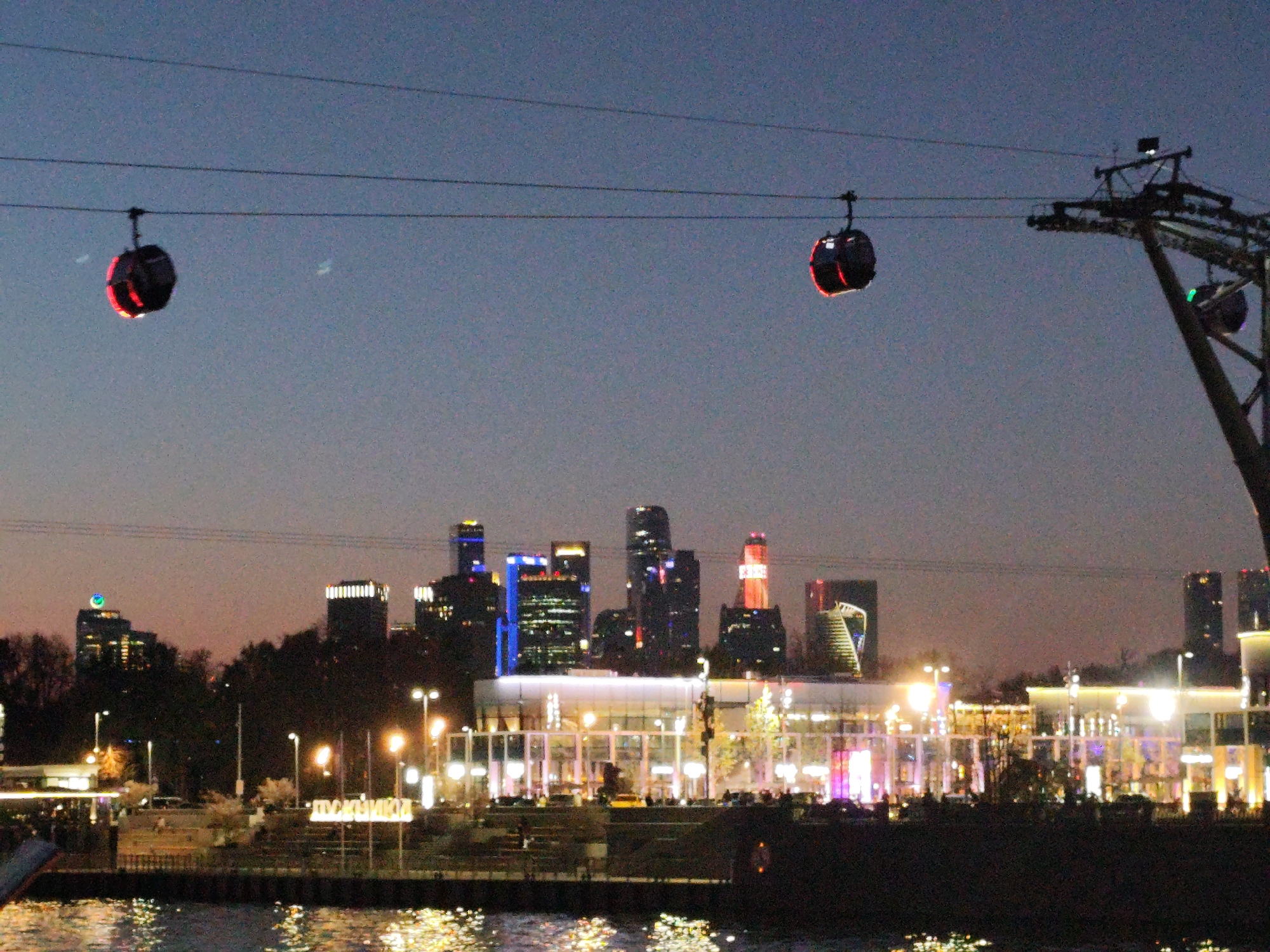 The image captures a city scene at dusk or night, with a body of water in the foreground reflecting the lights of the city. A cable car system is prominent, with gondolas suspended from overhead cables. The urban skyline features a cluster of skyscrapers with various architectural designs and illuminated windows. A brightly lit structure or complex is visible along the waterfront, with smaller lights and decorative elements. The sky is a gradient of dark blues and grays, and the overall scene gives the impression of a modern cityscape at night.