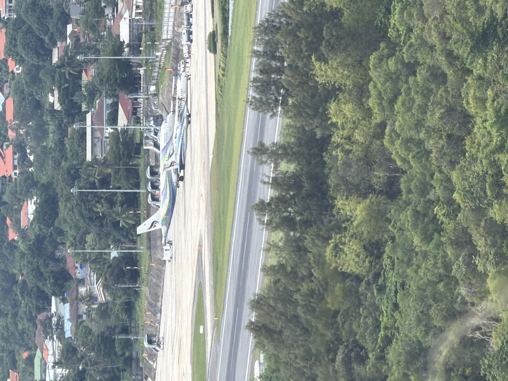 The image, captured from an elevated perspective, depicts an airport environment in Ban Bang Rak, Thailand. The foreground is dominated by lush green foliage, obscuring the lower portion of the scene. In the midground, an airport runway is visible, with two airplanes parked on the tarmac. The airplane on the right features a distinctive colorful livery. Several service vehicles and support equipment are present around the aircraft. Beyond the runway, a residential area with numerous houses, characterized by red and white roofs, is nestled amongst dense tropical trees that cover the surrounding hills. Several tall light poles are also visible across the airport grounds. The sky is overcast, contributing to the diffused lighting of the scene.