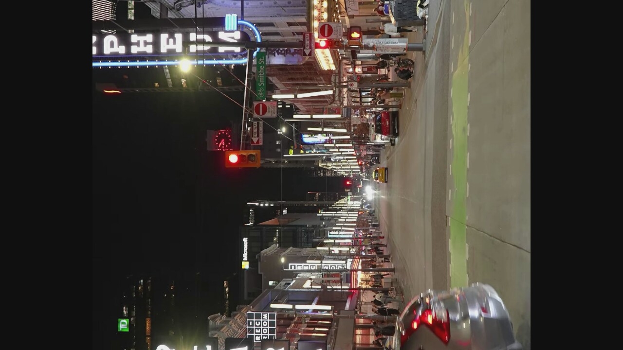 This is a night-time street scene in Vancouver, Canada, likely on Granville Street, known for its entertainment and theatre district. The image depicts a busy urban environment with a street lined with illuminated buildings and businesses. A traffic light is visible in the foreground, showing a red light. Several vehicles, including a yellow taxi and a red car, are present on the street. Pedestrians are seen walking on the sidewalks, with some interacting with each other. The architecture suggests a mix of older and modern buildings, many adorned with neon signs and vertical lighting, contributing to the vibrant nighttime atmosphere. The street appears to be a major thoroughfare, indicated by the presence of multiple lanes and traffic control devices.