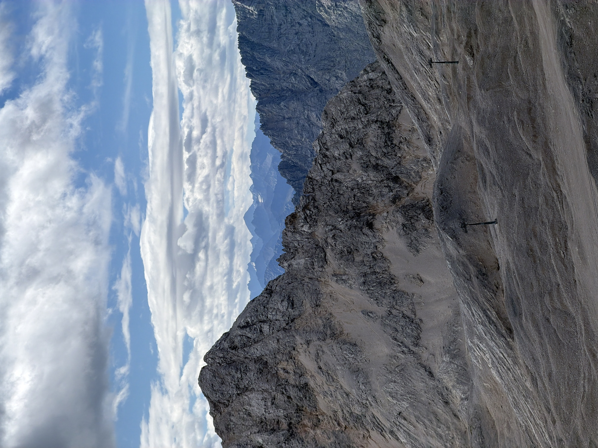 The image depicts a rugged, mountainous landscape under a partially cloudy sky. In the foreground, a barren, rocky terrain slopes downward, with two tall, dark, vertical poles, likely part of a ski lift or cable car system, visible on the ground. The mid-ground is dominated by a large, grey, rocky mountain face with textured surfaces and lighter colored scree or loose rock areas. Further in the background, a series of sharp, jagged mountain peaks, also appearing rocky and grey, recede into the distance, with some appearing dusted with snow. The sky is a mix of blue patches and white, fluffy clouds, with some darker, more ominous clouds also present. The scene is devoid of any people or visible wildlife. The overall impression is one of a high-altitude, natural environment, likely a ski resort or alpine region. Given the description of Garmisch-Partenkirchen, this is likely a view of the Zugspitze, Germany's highest peak, or the surrounding Wetterstein Alps.