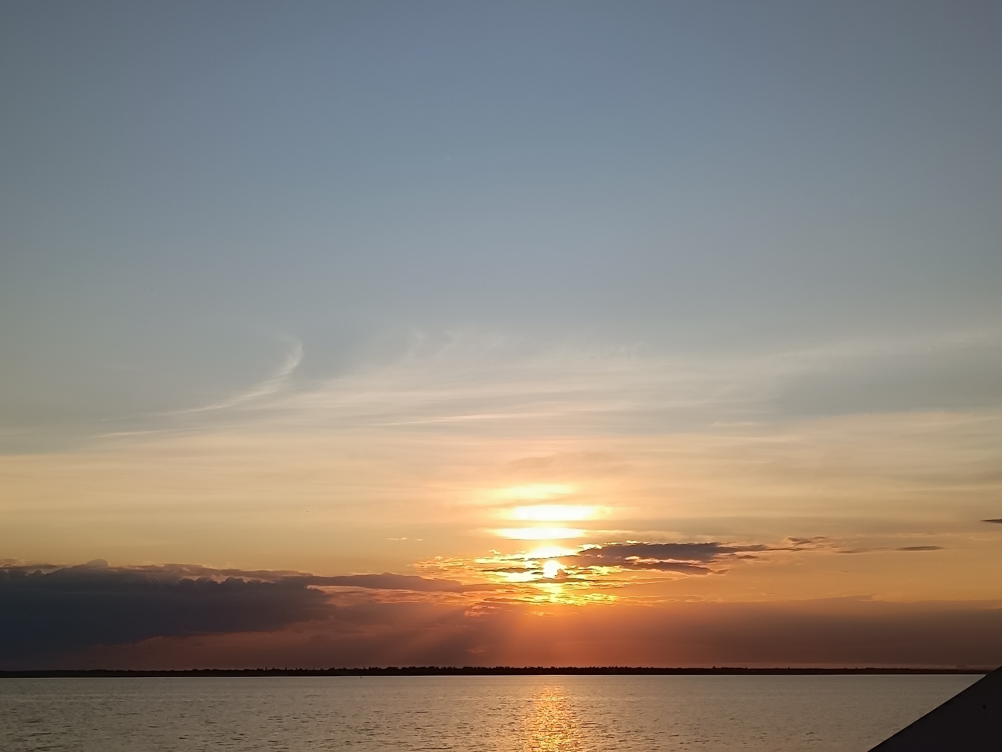 A tranquil sunset over a body of water is depicted in this image. The sky transitions from a pale blue at the top to warm hues of orange and yellow near the horizon, where the sun is setting. Rays of sunlight pierce through the clouds, creating a dramatic effect. The water below reflects the colors of the sky, with shimmering highlights. A dark, silhouetted landmass stretches across the horizon, suggesting a distant shore. A portion of a dark, angular object is visible in the lower right corner of the image, likely part of a structure or boat. The scene is serene and bathed in the warm glow of the setting sun.