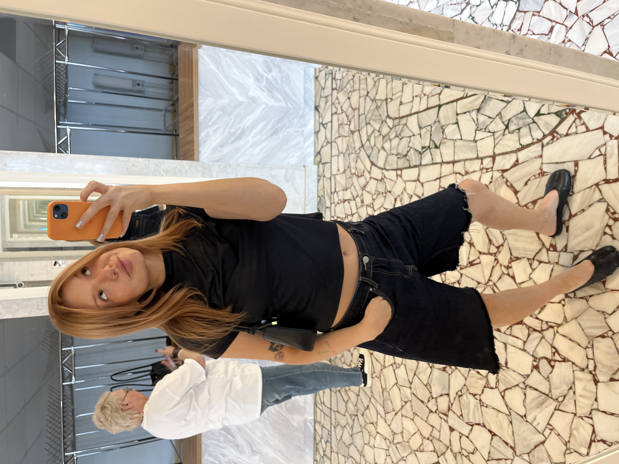 A young woman with auburn hair is taking a mirror selfie indoors, holding an orange smartphone in her right hand. She is wearing a black crop top, dark denim shorts with a frayed hem, and black split-toe flats. A small black shoulder bag is slung across her body. Behind her, a woman with short blonde hair, wearing a white t-shirt and blue jeans, is visible in the reflection of the mirror. The background suggests a commercial or retail space, possibly a boutique, with clothing racks and mirrors. The floor is a mosaic of light-colored tiles. The image does not contain any information that would place it in Chaikovsky, Russia.