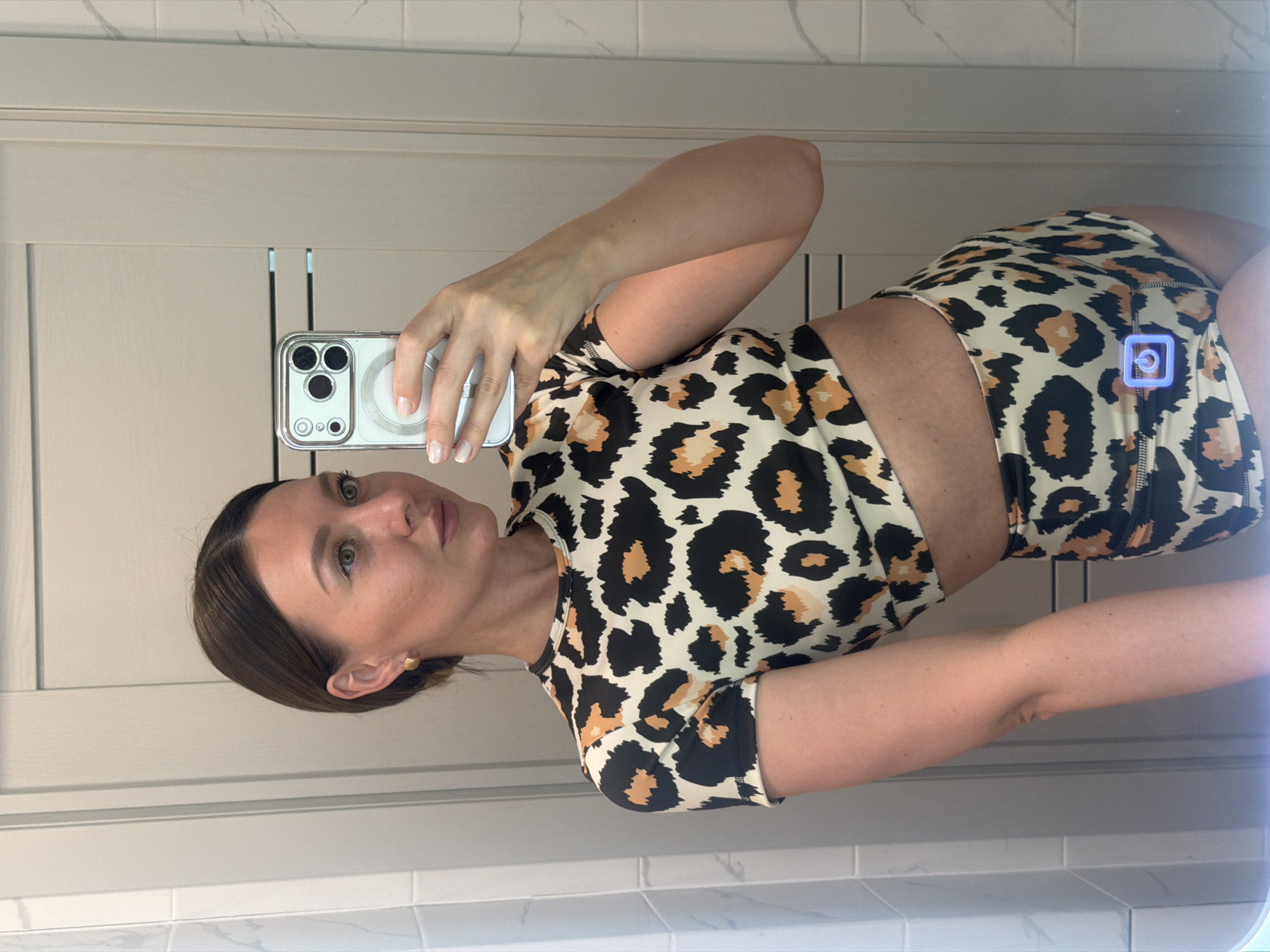 A woman is taking a selfie in a bathroom. She is wearing a leopard print athletic outfit consisting of a short-sleeved crop top and shorts. She is holding a smartphone in her left hand, with the camera facing her. The background shows a bathroom mirror and tiled walls. There is no evidence to suggest this image was taken in Rostov-on-Don, Russia.
