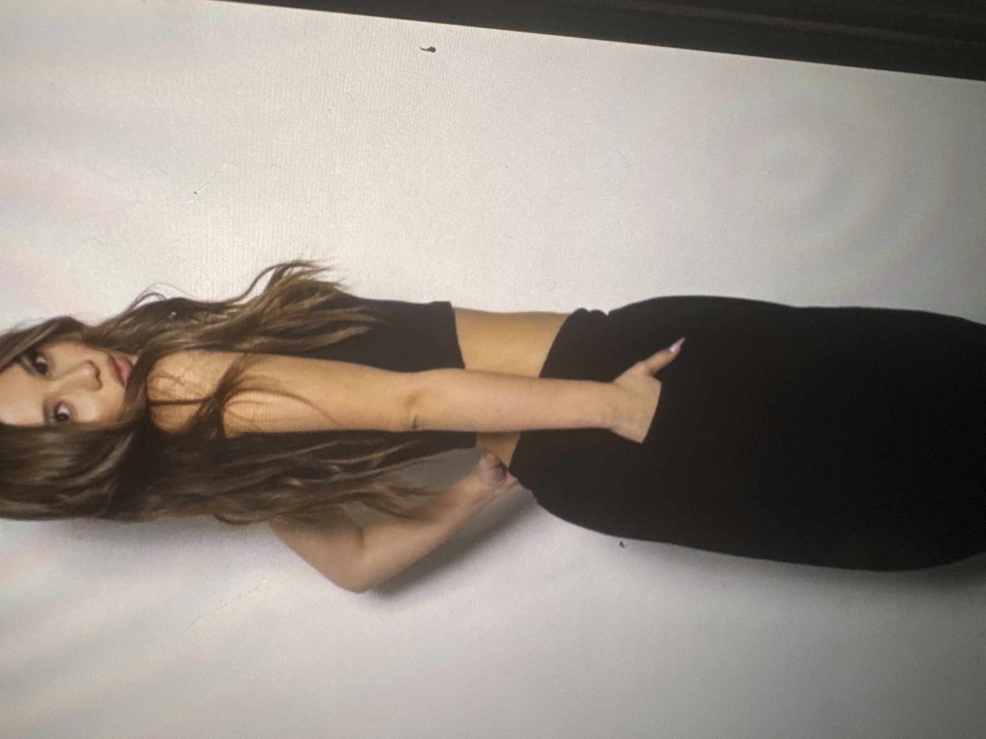 A full-body portrait captures a young woman posing against a plain, light-colored background. The image is oriented 90 degrees counter-clockwise, depicting her horizontally, though she appears to be standing upright in the original capture. She has long, wavy dark brown hair parted down the middle, flowing over her shoulders and chest.
She is dressed in a black crop top that reveals her midriff and high-waisted black bottoms, possibly pants or a skirt. Her left hand is casually tucked into her waistband or pocket, showcasing light pink or nude colored fingernails. Her right arm is bent at the elbow, resting near her side. The woman looks directly at the camera with a neutral, yet confident expression, her head tilted slightly.
The lighting is soft and even, suggesting an indoor studio or controlled environment. A dark band is visible at the very top of the image, likely indicating the edge of a backdrop or part of the studio setup. There are no specific location cues or visible text, and no distinct objects other than her attire. The overall mood conveyed is posed and composed.