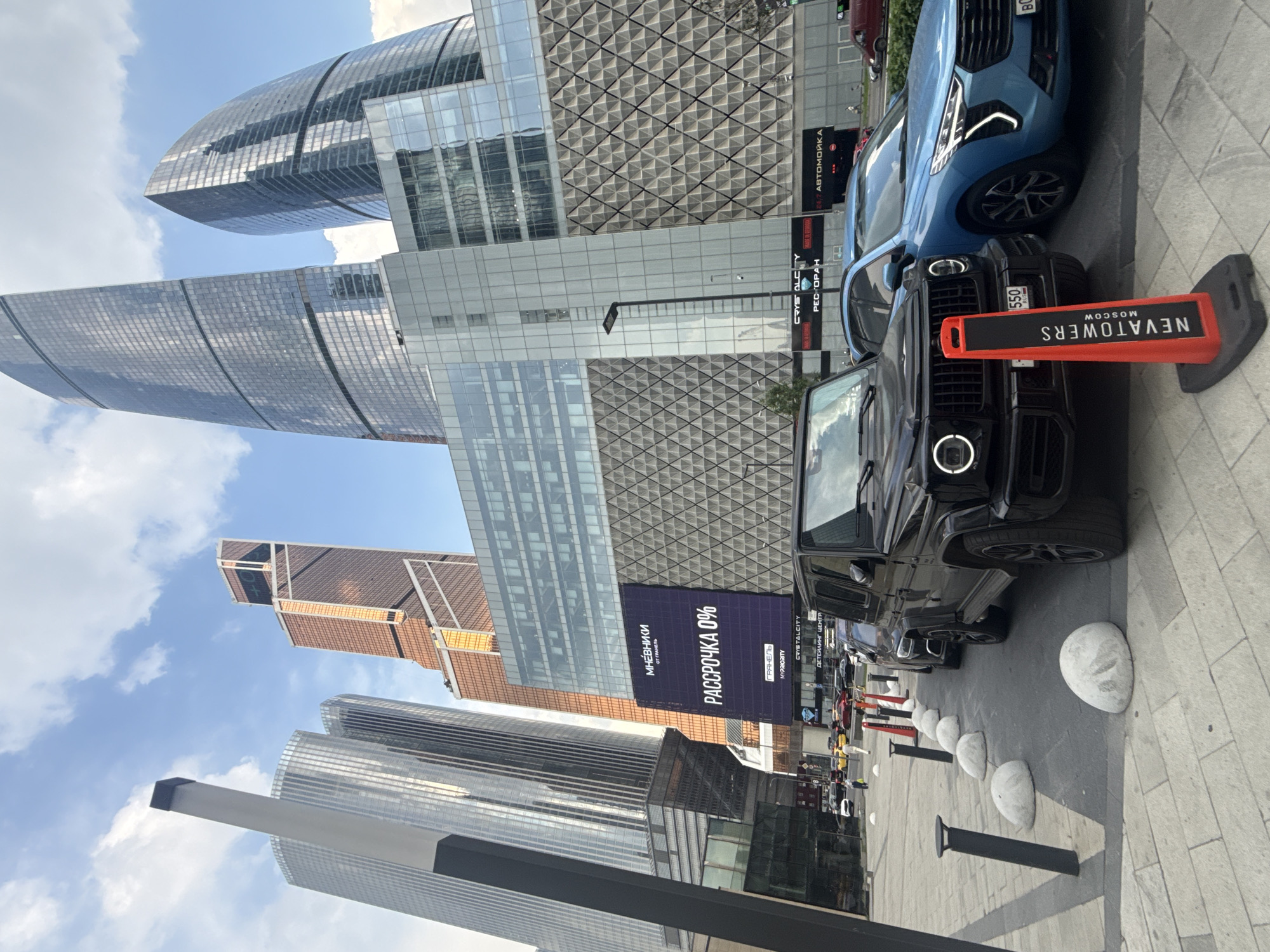 The image depicts a street view in the city of Khimki, Russia, specifically Moscow, as indicated by the "NEVATOOWERS MOSCOW" signage on an orange cone. In the foreground, a black luxury SUV, likely a Mercedes-Benz G-Wagon, is parked on the right, facing left. Next to it, a blue SUV is also parked. Several concrete bollards are lined up along the sidewalk, separating the pedestrian area from the street.
In the background, a large billboard displays text in Russian, prominently featuring "РАССРОЧКА 0%" which translates to "Installment 0%". Surrounding the billboard are modern skyscrapers with glass facades, characteristic of a metropolitan cityscape. The sky is partially cloudy. Traffic is visible on the street behind the parked cars, including yellow taxis and other vehicles. People are walking on the sidewalk in the mid-ground. The overall scene suggests a commercial district with high-rise architecture and active street life.
