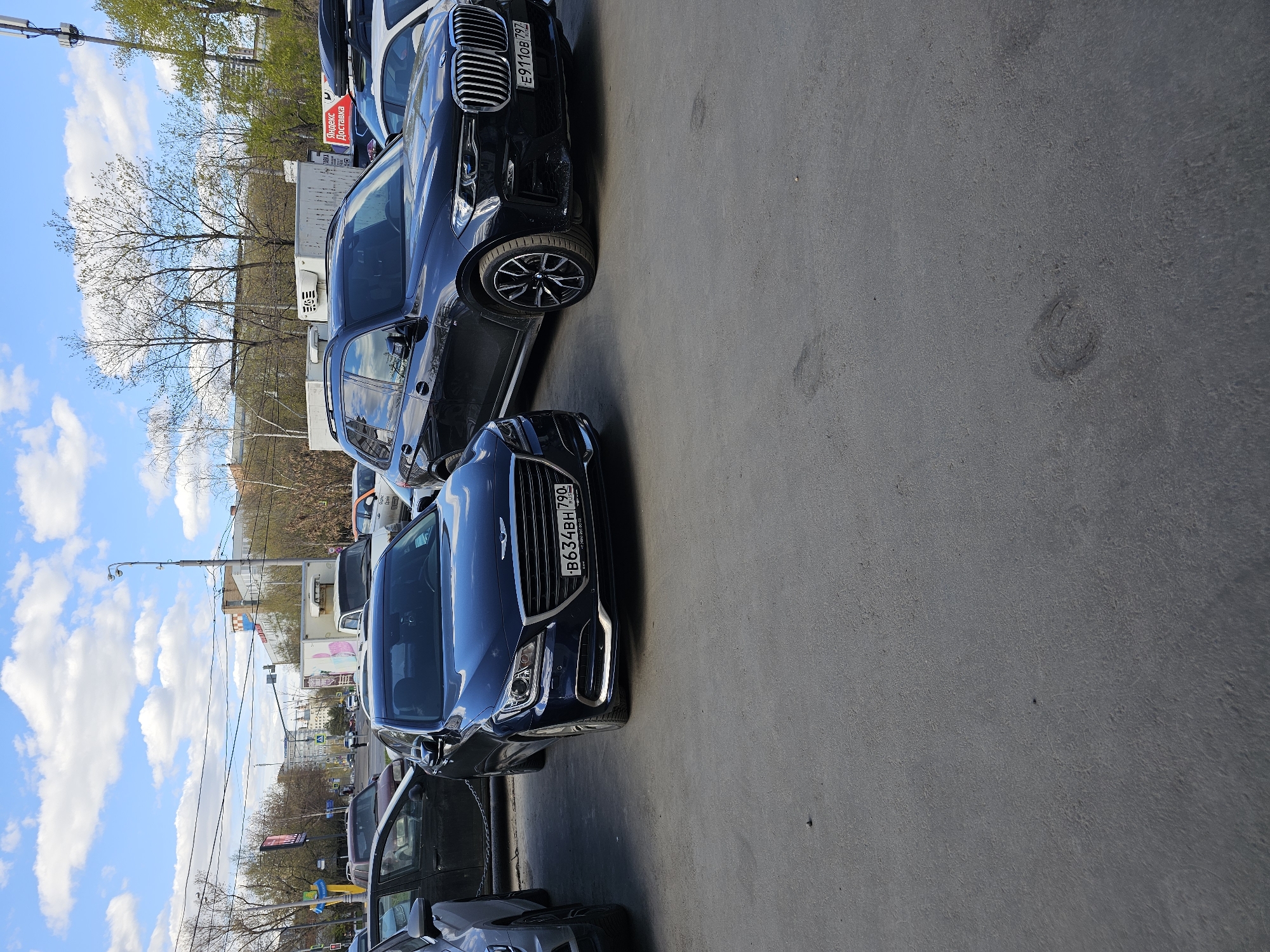 The media file presents a daytime street scene in Moscow, Russia. Several cars are parked along the right side of an asphalt road. In the immediate foreground, a dark blue Hyundai Genesis sedan is parked, with its front grille and headlights clearly visible. Behind it is a dark-colored BMW SUV, also parked. Further down the street, a white commercial van with a red "Яндекс Доставка" (Yandex Delivery) sign is visible.
The sky is bright blue with scattered white clouds, suggesting clear weather. On the left side of the image, the upper branches of bare trees are visible, with some hints of budding leaves. Buildings and utility poles line the street in the background, characteristic of an urban environment.
Visible text includes the Russian license plates on the parked cars: "B634BH 790" on the Genesis sedan and "E911OB 797" on the BMW SUV. Both region codes (790, 797) correspond to Moscow and Moscow Oblast. The "Яндекс Доставка" text on the white van is also prominent. The scene is quiet, with no people actively present, focusing primarily on the parked vehicles and the street environment.
