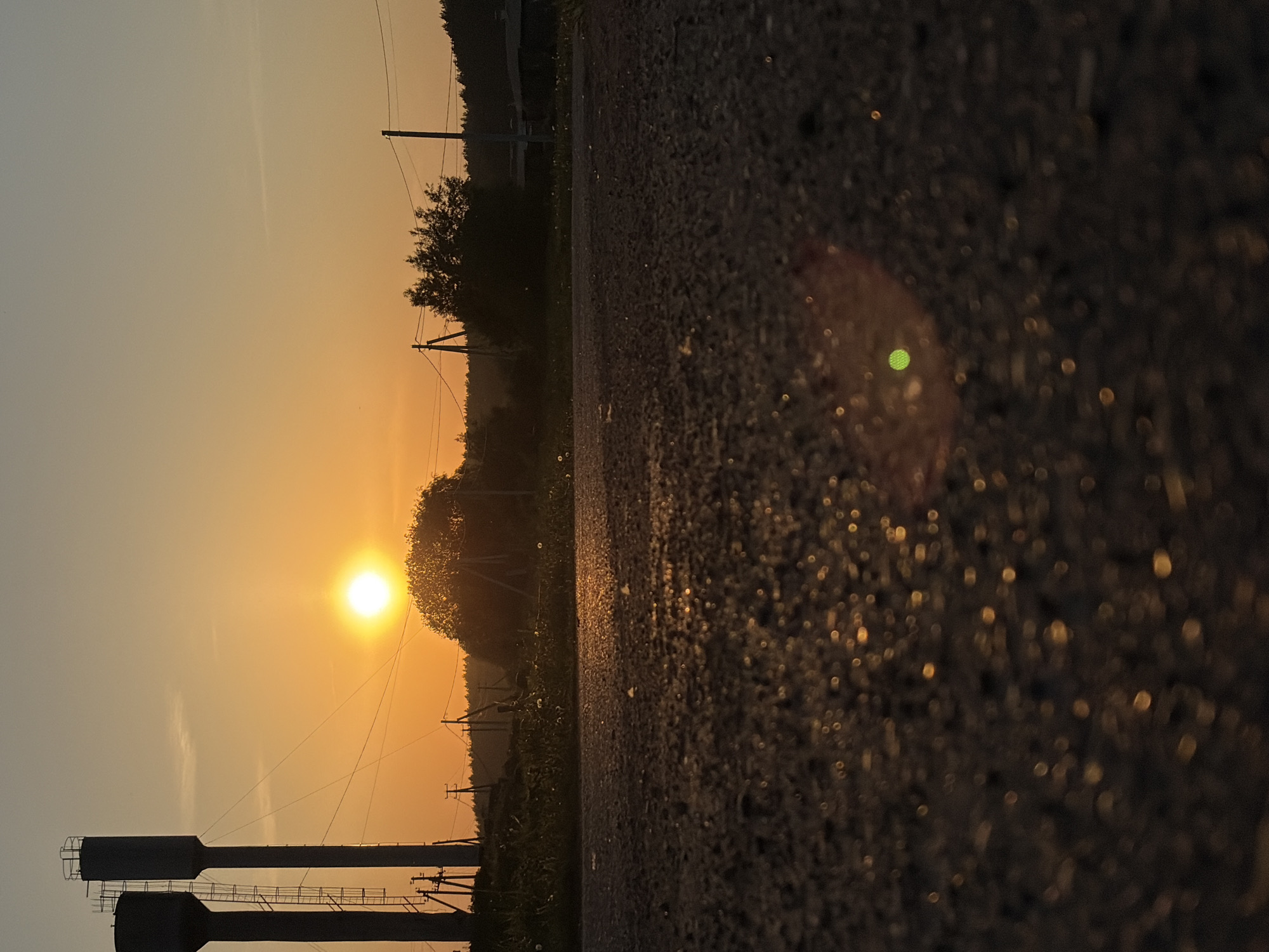 The image depicts a low-angle view of an asphalt surface, likely a road, in the vicinity of Чумна, Russia, during a sunset. The sun is prominently featured in the sky, casting a warm, orange glow across the scene. Silhouettes of trees, vegetation, and power lines are visible against the bright sky. In the foreground, there are two water towers or similar industrial structures. The asphalt surface is the dominant feature of the foreground, displaying granular texture and reflective points of light.