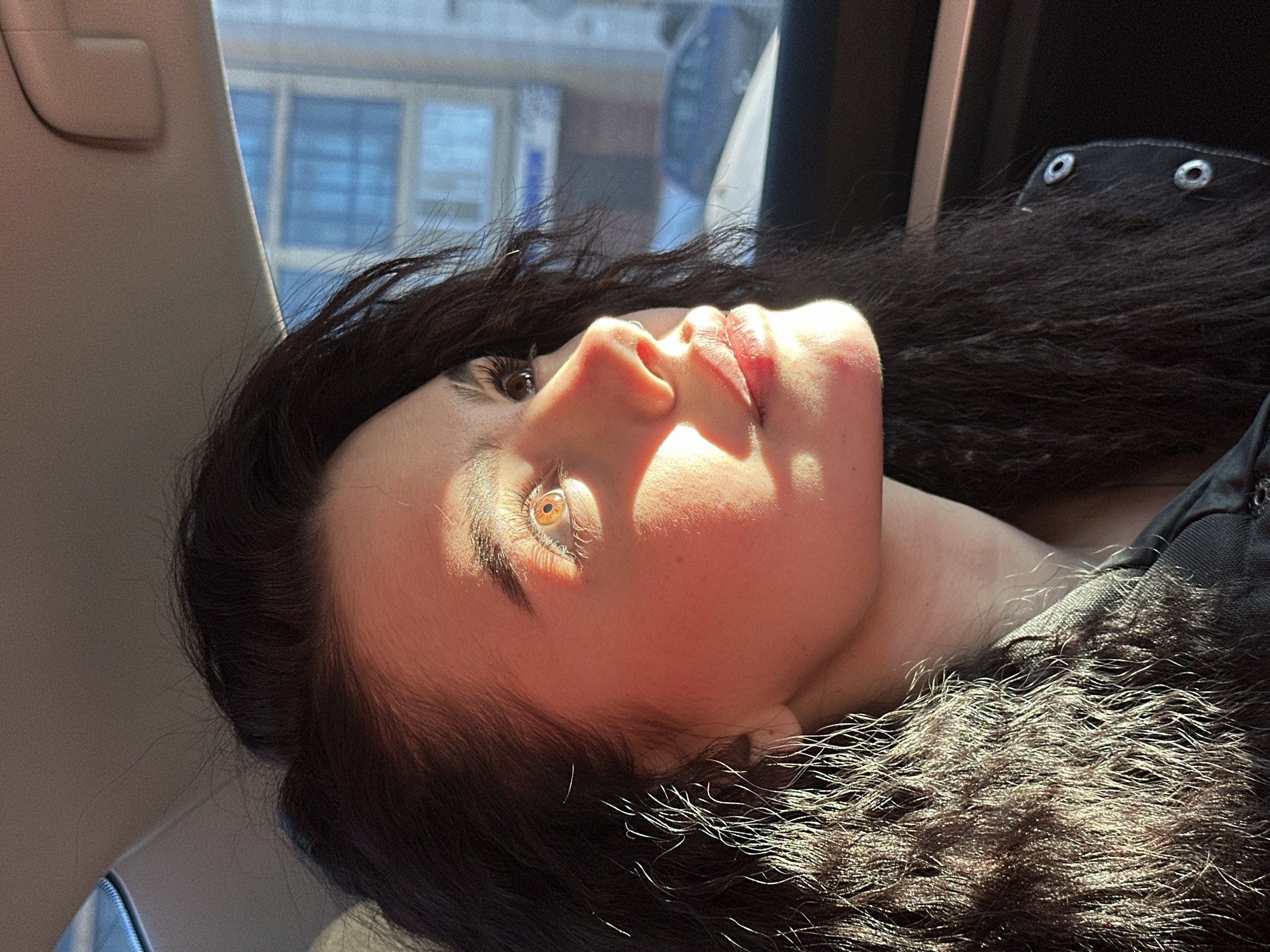 A close-up, angled portrait shows a woman lying down inside a vehicle, possibly a car. The interior of the vehicle features a tan-colored pillar and what appears to be the edge of a window with a blurred urban scene visible outside. The woman has dark hair and fair skin, with sunlight highlighting her face. She is wearing a dark-colored garment with visible metal snap details. Her eyes are open, and she is gazing upward. The sunlight is casting strong shadows on her face, creating a high-contrast image. There is no clear indication of the location being Kazan, Russia based on visual elements.