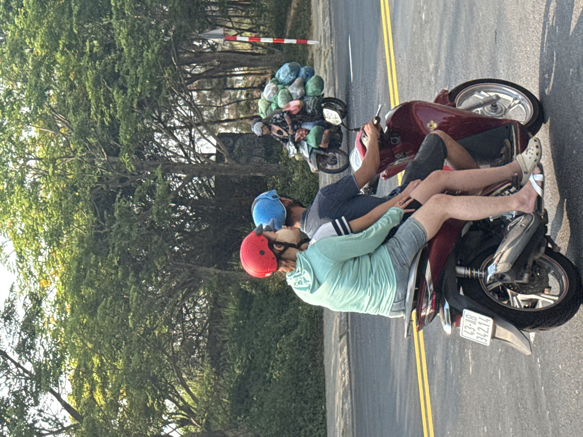 Two motorcycles are on an asphalt road with double yellow lines. A red and white safety barrier is visible in the background. The first motorcycle carries a large bundle of bags. On the second motorcycle, two people wearing helmets ride. The driver is wearing a blue helmet and the passenger is wearing a red helmet. The license plate of the second motorcycle reads "43-AB 342.14". Lush green trees line the side of the road. The location is Đà Nẵng, Vietnam.