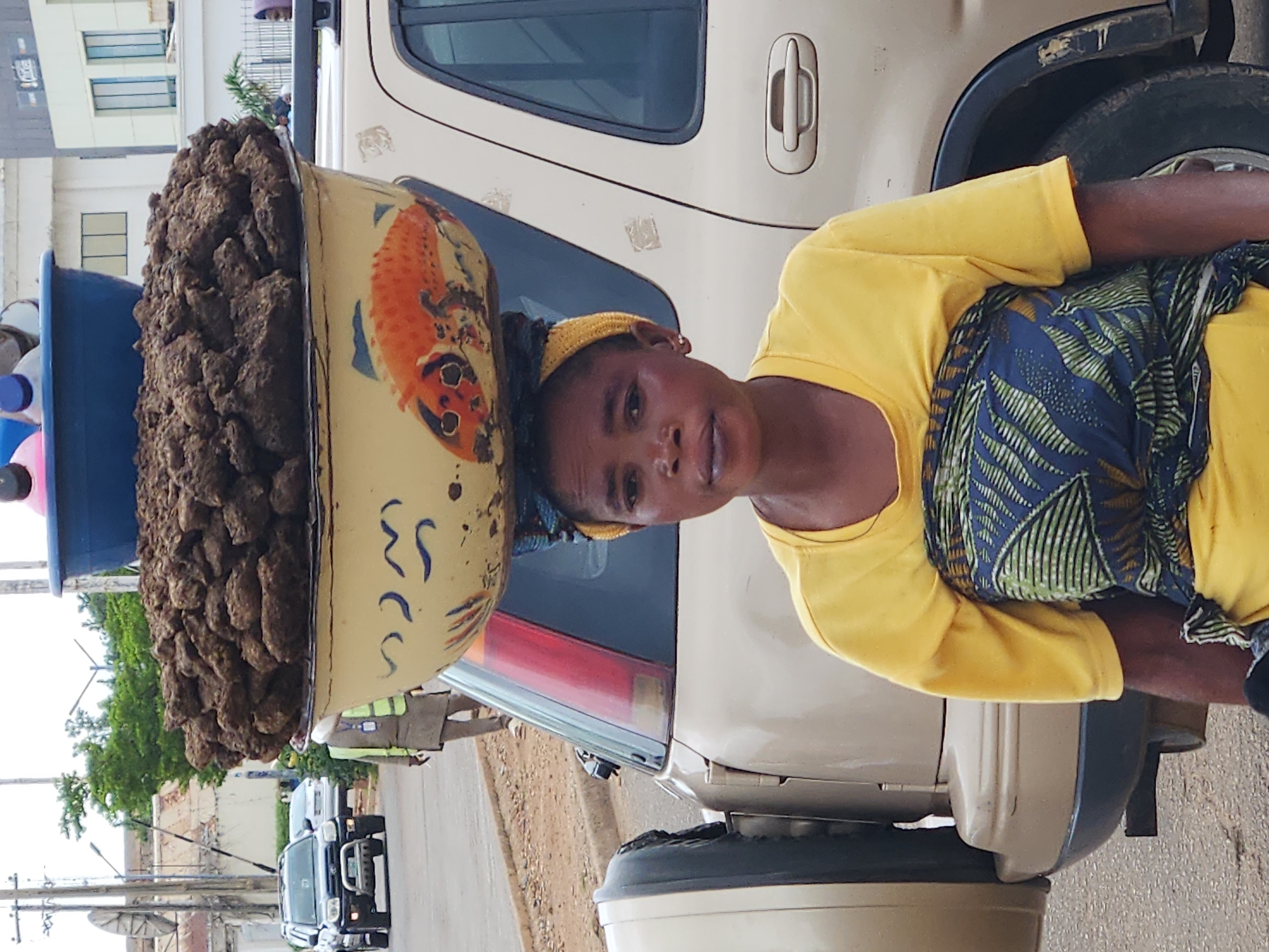 In the city of Jos, Nigeria, a woman carries a large bowl of what appears to be dried animal dung on her head, balanced on top of a blue plastic basin. The bowl itself is yellow and decorated with a painted orange fish. She is wearing a yellow t-shirt and a patterned fabric wrapped around her lower body. Behind her, a tan SUV is parked, with its spare tire visible on the rear. In the background, other vehicles and buildings suggest an urban setting. The ground is paved, and there is some sparse vegetation visible.