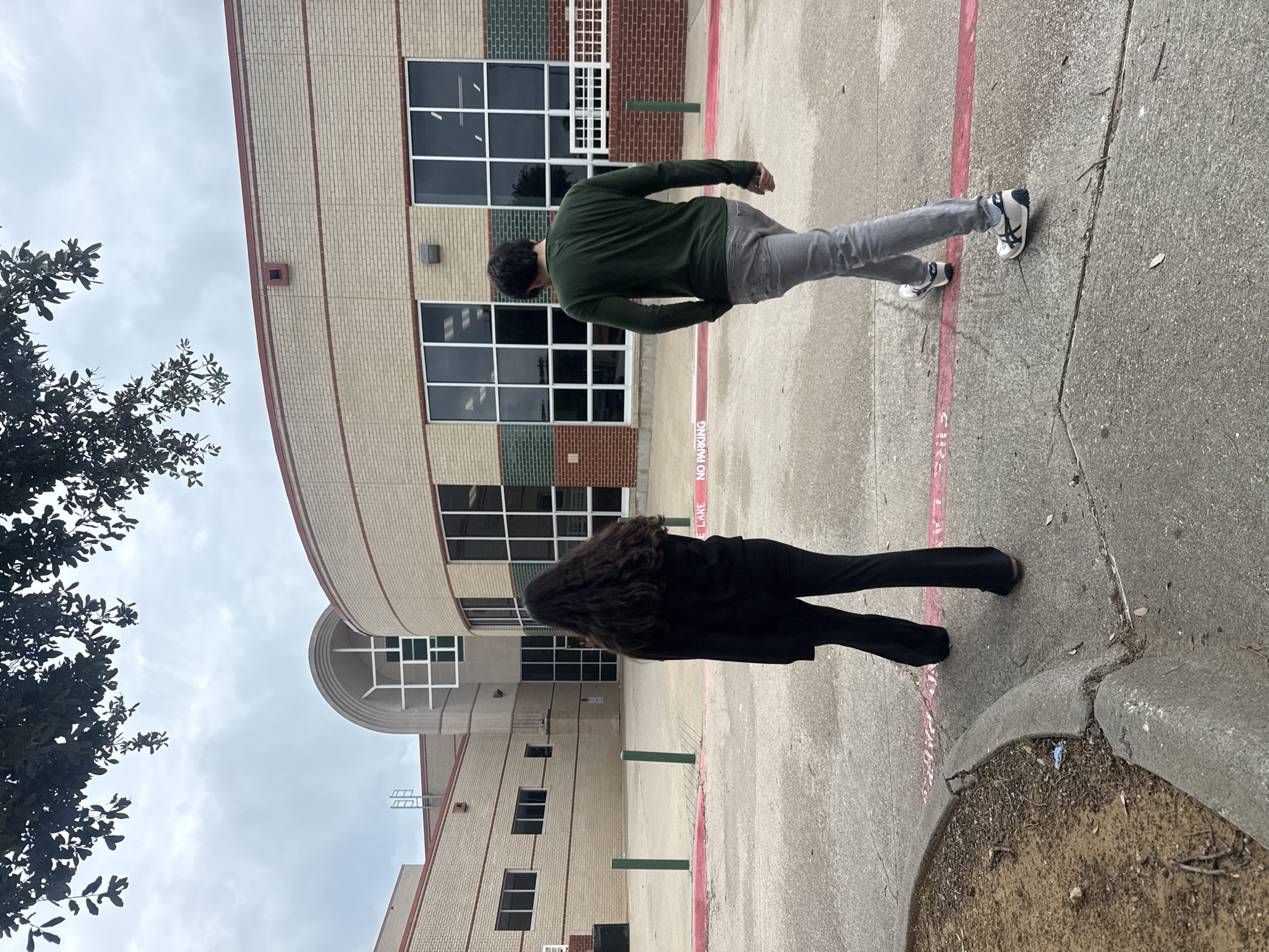 The image depicts two individuals walking on a concrete surface adjacent to a brick building.
One person is wearing a dark green long-sleeved shirt and gray pants, paired with sneakers that appear to have a white body and black accents. The other person is wearing an all-black outfit. Both individuals are walking away from the camera, seemingly along a red painted line that reads "NO PARKING" on the surface.
The building behind them is predominantly brick, with a combination of lighter tan bricks and darker green sections. The structure features several windows with white frames. The sky is overcast. A tree with dark foliage is visible to the side. The scene is located in Southlake, United States of America.