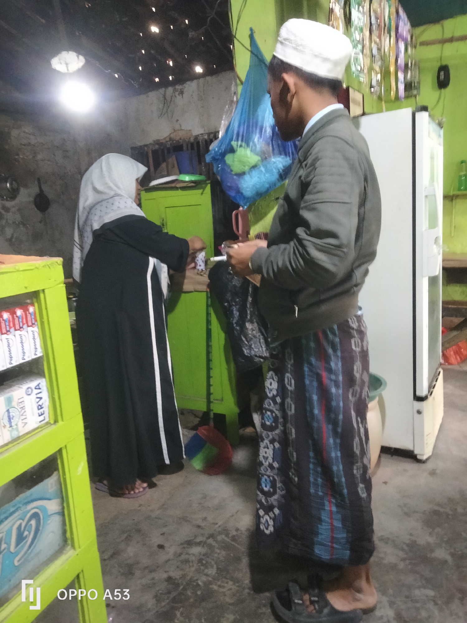 The image depicts an indoor scene, likely a kitchen or storage area, with two individuals. A woman wearing a hijab and a black abaya stands to the left, facing a green cabinet. She appears to be reaching into the cabinet. To her right, a man wearing a white peci cap and a dark jacket over a sarong stands facing away from the camera. He is holding a cigarette or a similar object in his right hand and looking towards the woman. In the background, a white refrigerator is visible. The environment appears somewhat cluttered, with a bright overhead light illuminating the space. Shelves with boxes of "Pepsodent" toothpaste and "Lervia" soap are visible on the left side of the frame. A plastic bag hangs in the background. The floor is concrete. The OPPO A53 logo is present in the bottom left corner. The scene is consistent with a domestic setting in a moderately lit area in Badung, Indonesia.