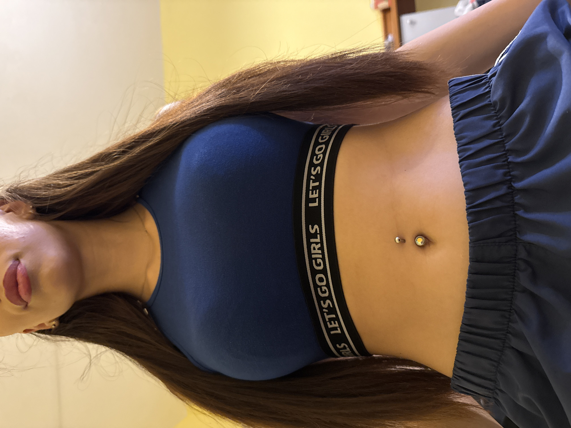 A low-angle, medium shot captures a woman from the waist up, wearing a dark blue sports bra with "LET'S GO GIRLS" printed on a black band and dark blue athletic shorts. Her long, dark brown hair frames her face, which is partially visible with her lips slightly parted and wearing light pink lipstick. She has a navel piercing with a silver bar and a yellow gem. The background is a plain, light-colored wall with a yellow tint on the right side, suggesting an indoor setting. The lighting is warm and appears to come from the right.