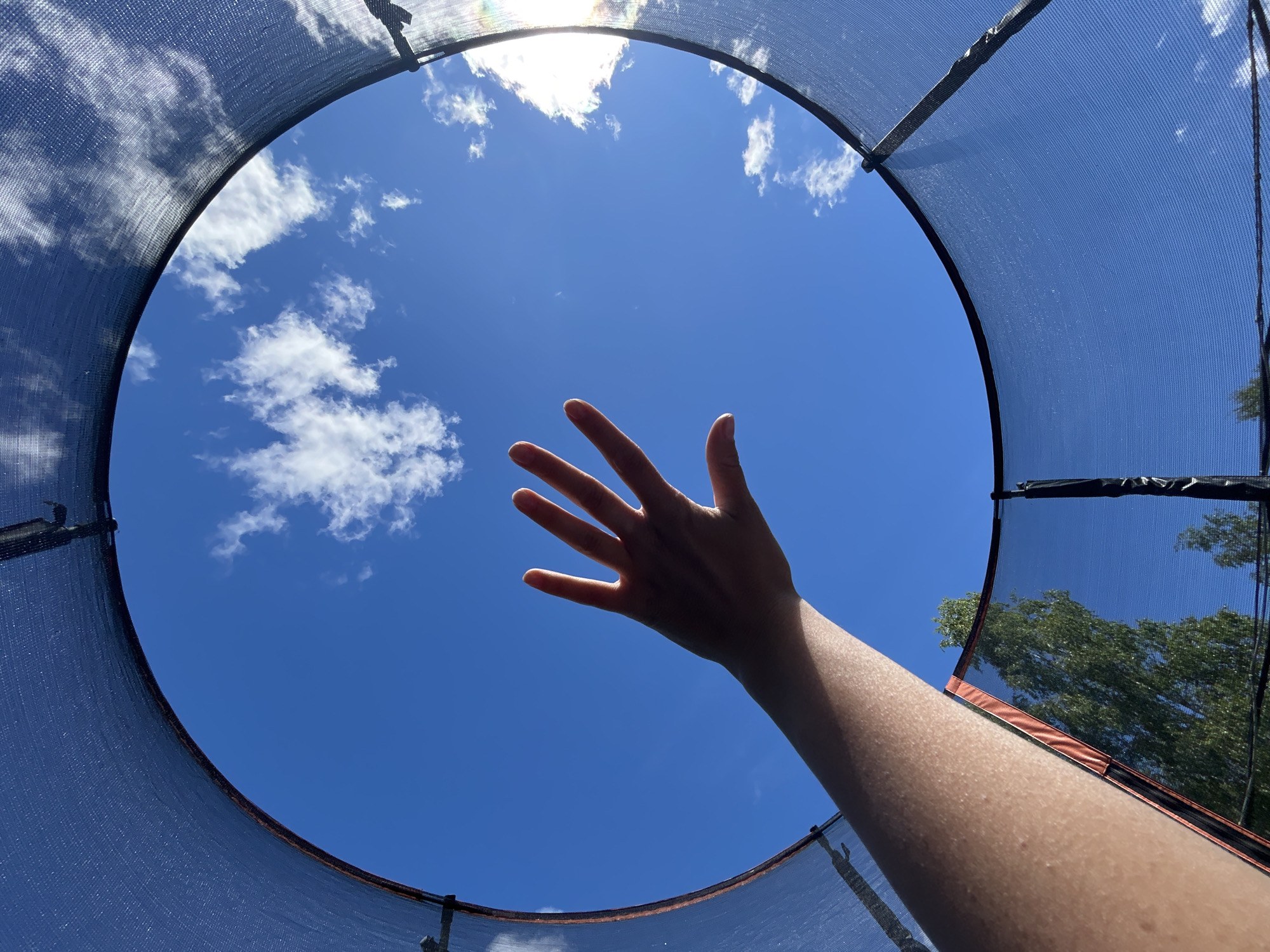 An arm and hand are reaching upwards towards a bright blue sky filled with scattered white clouds. The viewpoint is from inside a trampoline, looking out through the safety net. The edge of the trampoline's dark netting curves around the frame, with hints of orange padding visible underneath. Trees can be seen in the lower left and right corners of the frame, suggesting an outdoor setting, likely a backyard in Novozhilovo, Russia. The lighting indicates a sunny day.