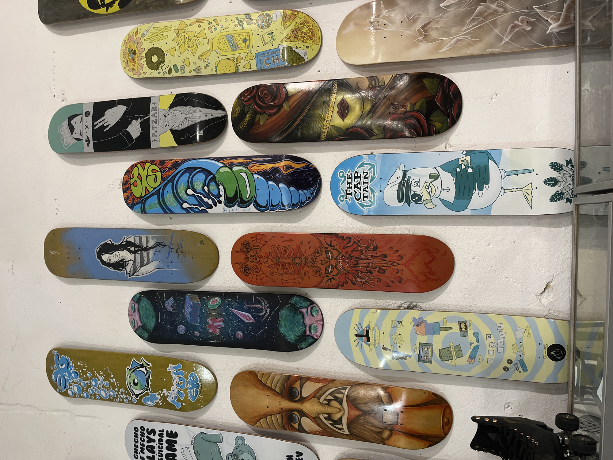 The image displays a collection of 14 skateboards mounted on a white wall. Each skateboard features unique artwork, ranging from cartoonish characters and abstract designs to realistic portraits. Some skateboards have text, including the words "CHEESE," "FREE WIN," "P.TZAR," "SURVIVAL," "THE CAPTAIN," "SUICIDAL," "AME," "CHECHO," and "EMECHO." The skateboards are arranged in a staggered, grid-like pattern. The lighting is bright and even. A portion of a mirrored cabinet is visible on the right side of the image. Black roller skates are present in the bottom-right corner of the frame. Based on the text on one of the skateboards, the skateboards are of Checho Lays Suicidal AME. There are no people visible in the image. There is no visual information in the image to indicate the city or location.