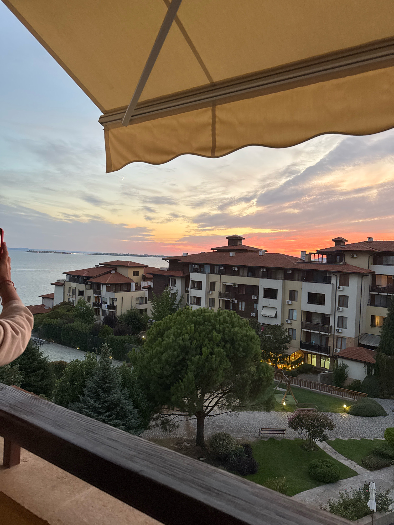This is a photograph taken from a balcony in Sveti Vlas, Bulgaria, during a sunset. A beige retractable awning is visible in the upper portion of the image. Below the awning, the sky displays a gradient of colors, with hues of orange, pink, and purple, indicating the sun is setting over the sea. The sea itself is visible in the background, with a distant coastline and scattered lights. In the middle ground, several multi-story apartment buildings with red-tiled roofs are clustered together. Green trees and shrubs are scattered throughout the scene, with a playground visible in the courtyard of one of the buildings. A person's arm and shoulder are visible on the left side of the frame, holding a red mobile phone, as if taking a photo. The foreground of the image is occupied by the wooden railing of the balcony.