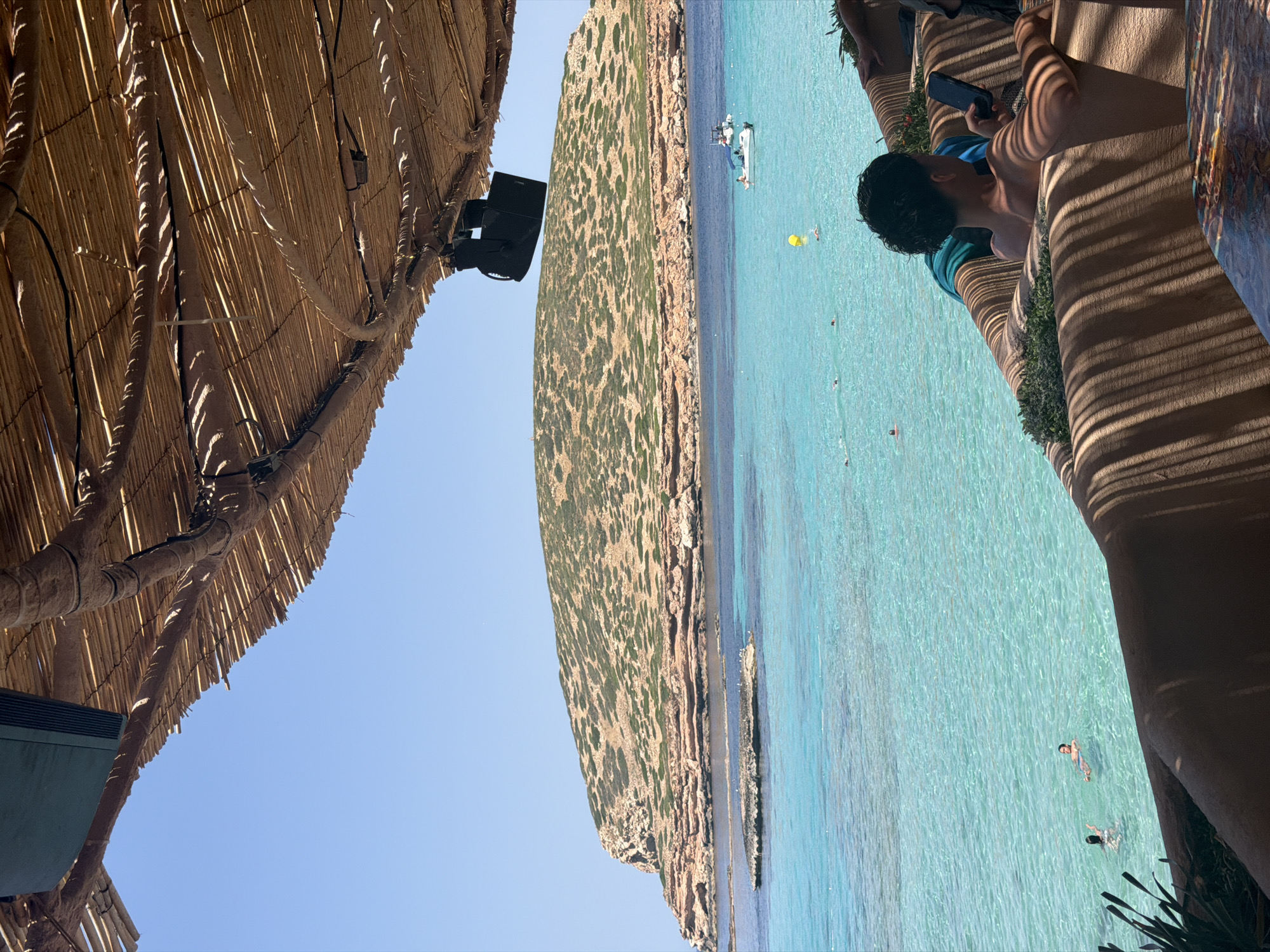 This image shows a view of a beach with clear turquoise water and a rocky, vegetated cliff face. The scene is viewed from under a thatched roof structure, with part of the roof and a speaker visible in the foreground. In the water, there are several people swimming, a yellow floating object, and a small white boat. The sun is shining brightly, suggesting a warm, clear day.
The perspective of the photo is tilted, making it appear as if the beach and sea are on a side plane. In the right side of the image, a person is sitting on a bench-like structure, looking at a phone. Their body is partially in shadow cast by the thatched roof, creating a striped pattern on the bench. The overall atmosphere appears relaxed and leisurely, typical of a beach vacation. The location is identified as Sant Josep de sa Talaia, Spain, which is known for its beautiful beaches. There is no visible text in the image.