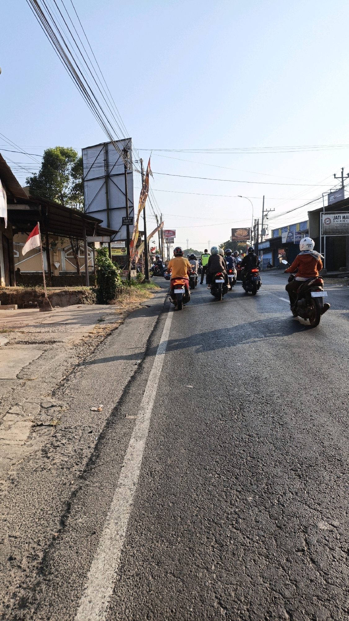 The image shows a street scene in Kuwasen, Indonesia, with a clear blue sky and bright sunlight. The road is asphalt, with a white line marking the lane. Several motorcycles are on the road, most carrying one or two riders. The motorcycles are of various models and colors.
On the left side of the road, there are buildings with a tiled roof and a sign that appears to be an Indonesian flag. There is also a large billboard with a white surface. Further down the road, on the right, there are more buildings and signs, one of which reads "JATI MAJU FURNITURE."
The people on the motorcycles are wearing helmets and casual clothing. Some are riding alone, while others have passengers. The overall impression is of a typical day on a road in a populated area, with a moderate flow of motorcycle traffic. The environment appears to be a mix of commercial and residential buildings along the road.