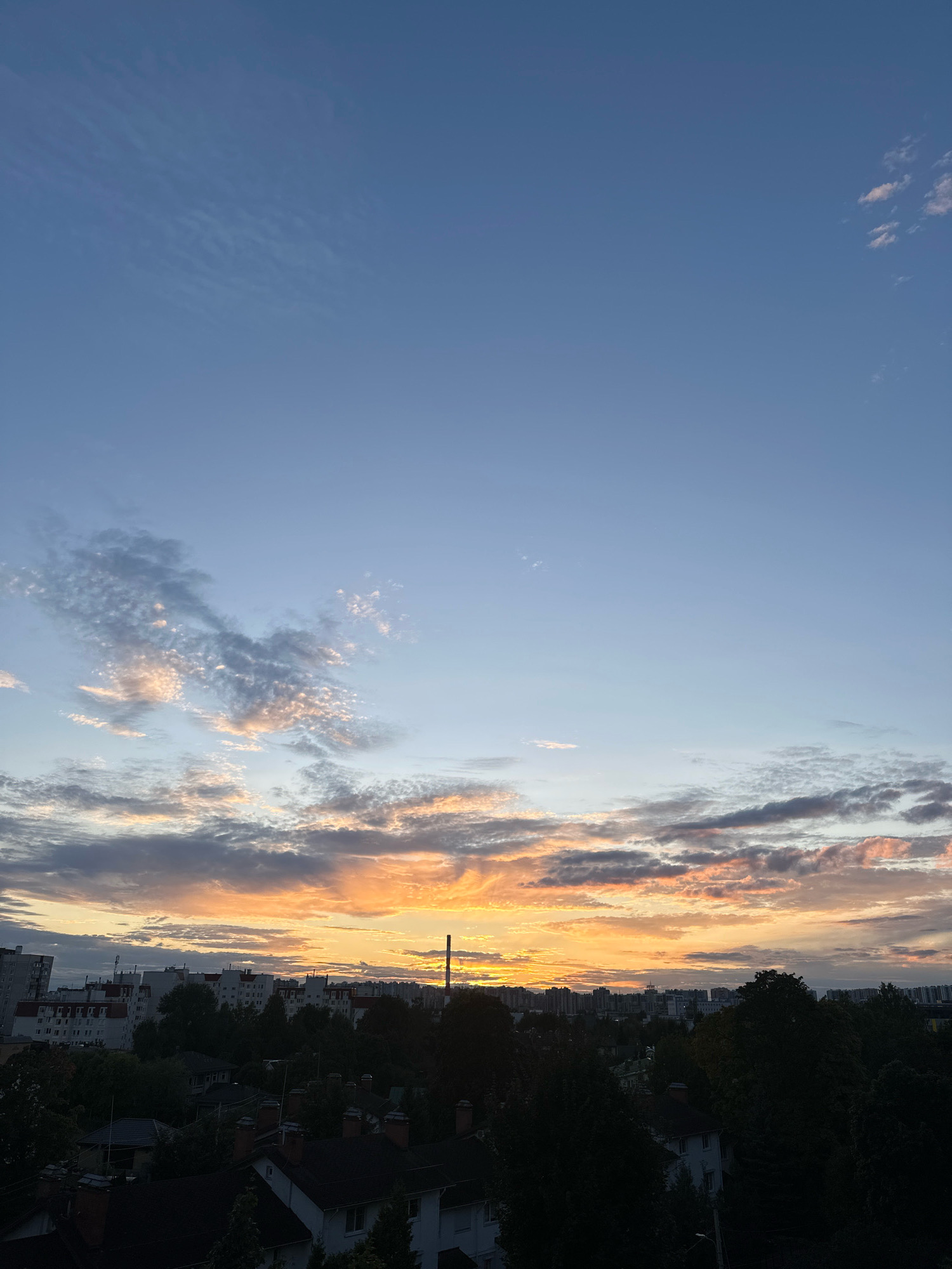 The image shows a city skyline at sunset. The sky is a gradient of blue to orange, with clouds illuminated by the setting sun. Below the sky is a dense arrangement of trees, interspersed with the rooftops of houses and apartment buildings. A tall, slender smokestack or industrial chimney is visible in the middle distance, rising above the trees and buildings. The overall impression is one of an urban environment at dusk, with natural elements of trees and sky interacting with man-made structures.