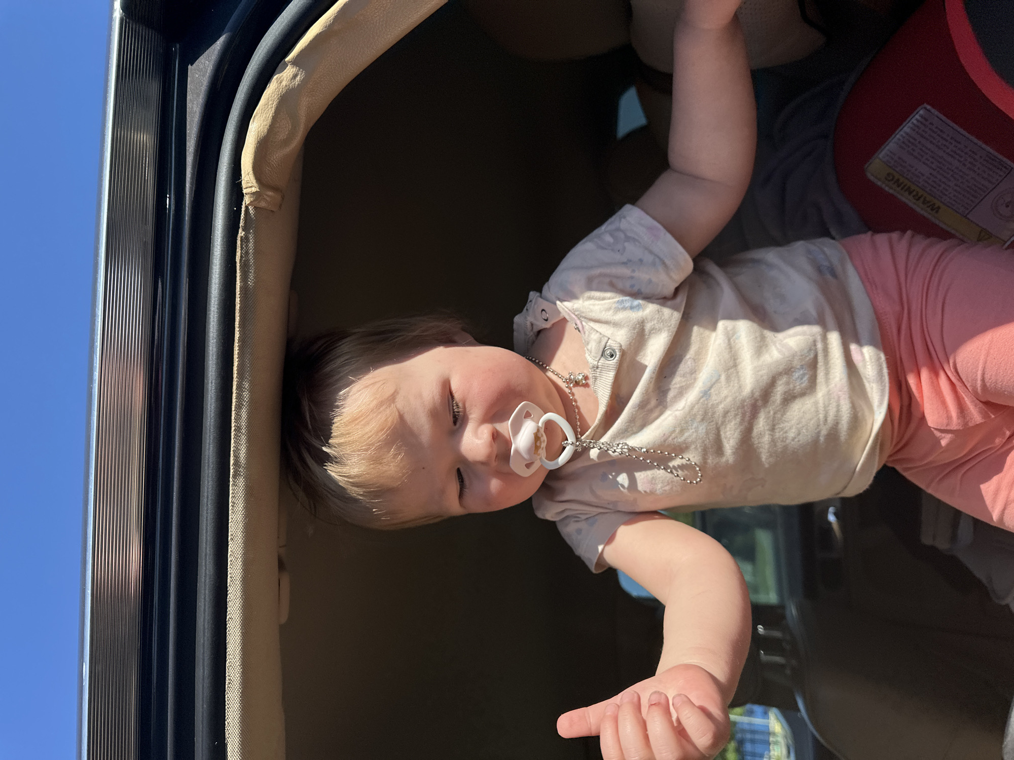 A young child, appearing to be a toddler with fair skin and short blonde hair, is positioned upright within a vehicle's opening, possibly a trunk or open window. The child is wearing a light-colored, short-sleeved t-shirt with faint patterns and pink leggings. A white pacifier with a heart-shaped shield and a silver-colored beaded chain is in the child's mouth. A thin, silver-colored chain necklace with a small cross pendant is visible around the child's neck. The child's left hand is raised, extended forward with the thumb pointed upwards.
The immediate environment consists of the car's interior, showing a light beige fabric headliner, dark rubber seals, and plastic trim around the opening. Above these, a horizontal metallic trim piece, possibly chrome or polished aluminum, is visible. In the lower right foreground, a red child safety seat is partially visible, displaying a yellow "WARNING" label. A clear, bright blue sky is visible above the car opening, indicating daylight conditions. Strong direct sunlight illuminates the child and parts of the car's interior, creating shadows within the vehicle. There are no discernible visual cues within the image that would allow for the geo-localization of the scene to Nizhny Novgorod, Russia.
