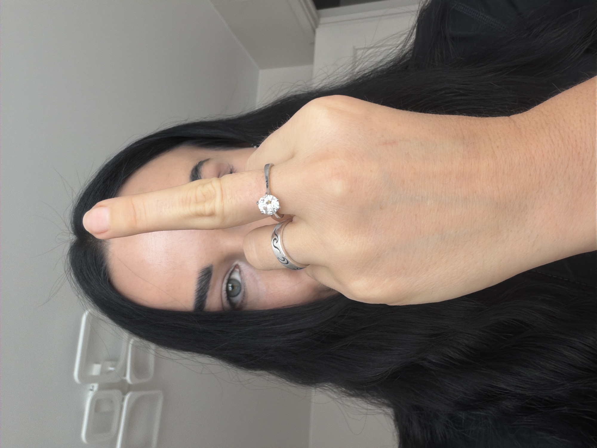 An upright, close-up shot features a person, likely female, with long, dark hair, partially obscuring their face with their hand. The person's left eye and eyebrow are visible, revealing a greyish-blue iris and well-defined dark eyebrows. The hand, positioned with the ring finger extended horizontally across the face, draws attention to two rings on that finger. The top ring is a silver-toned band featuring a prominent, round, clear gemstone, possibly a diamond. Below it is another silver-toned band with a patterned or wavy design.
The person is wearing a dark, possibly black, garment. The background is a plain, light-grey wall, with some indistinct white, rectangular shapes visible in the lower left corner, hinting at either shelving or structural elements. The gesture of the extended ring finger in front of the face could imply a variety of interpretations, from a dismissive or defiant stance to simply resting the hand. No specific time of day, weather, or location cues are discernible from the image itself, beyond being indoors. There is no visible text.