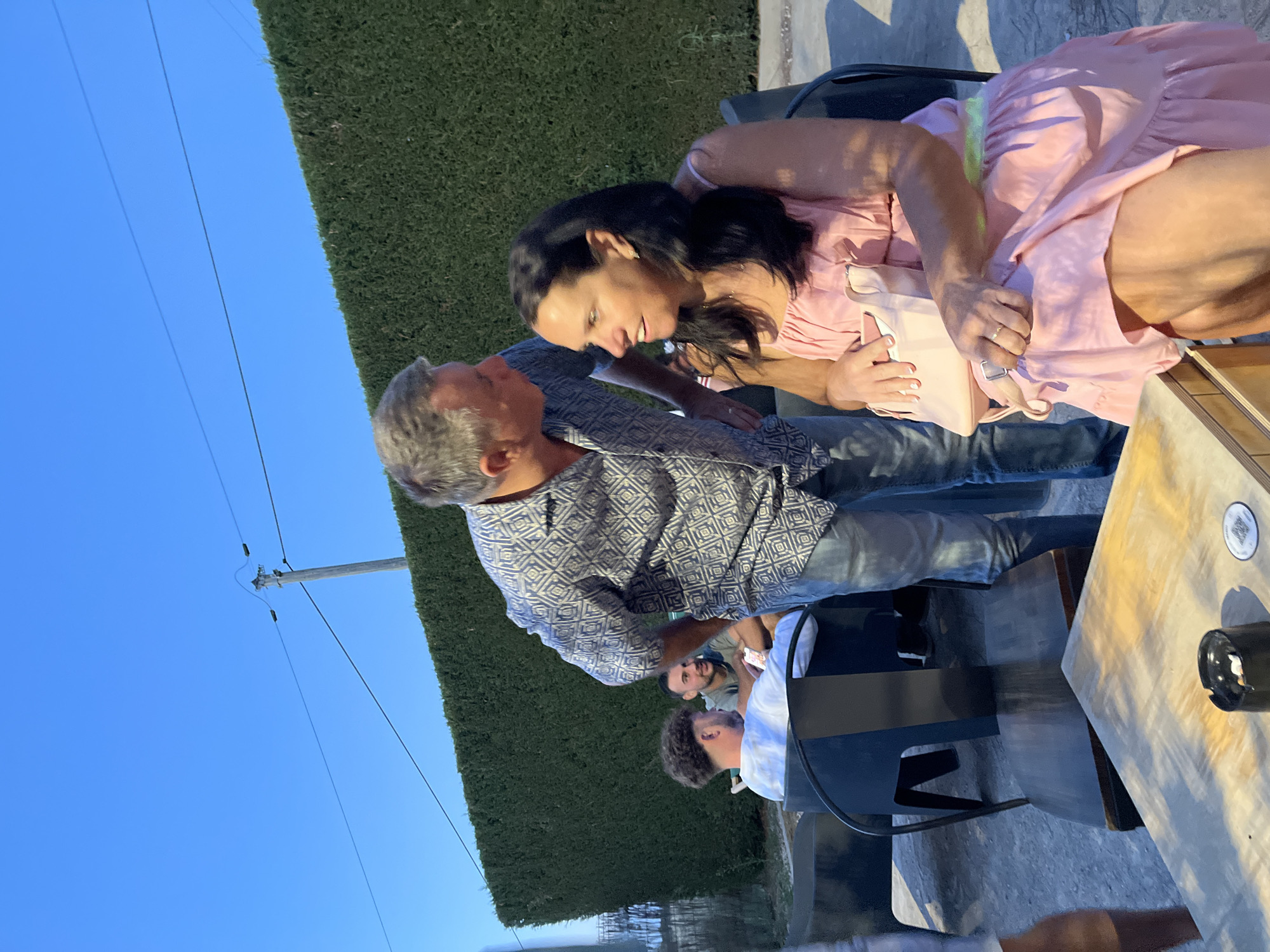 In Savigneux, France, a man in a patterned blue and white shirt leans in to speak with a woman wearing a light pink dress, seated outdoors. The woman holds a light pink purse on her lap, and a ring is visible on her right hand. Behind them, a dense green hedge forms a backdrop. In the background, two other men are seated, one with curly hair and another with short dark hair, both appearing relaxed. To the left of the frame, a wooden table with an ashtray containing cigarette butts and a QR code sticker is visible. The scene is illuminated by natural daylight, with a clear blue sky above.