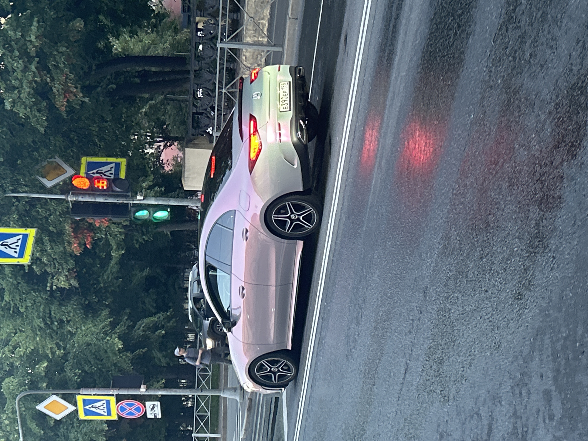 The image shows a side view of a car parked on a wet road. The car is a light-colored sedan with a pearlescent finish, reflecting the surroundings. Its taillights are illuminated red, casting a glow on the road. The road itself appears damp, with white lane markings visible. In the background, lush green trees form a dense canopy, suggesting an outdoor setting possibly a city street or a parkway.
Traffic lights are visible, with one displaying a red light and another showing green. Several road signs are also present: a yellow diamond-shaped sign, a blue rectangular sign with a pedestrian crossing symbol and an arrow, and a circular sign with a red border and a blue background indicating a prohibition. A license plate on the car reads "E390EP 198".
A person in casual clothing, wearing a cap, is visible on the sidewalk, partially obscured by trees. The overall lighting suggests it might be late afternoon or early evening, given the presence of illuminated traffic lights and the subdued ambient light. The wet road indicates recent or ongoing rain, contributing to a slightly muted and reflective atmosphere. The scene appears to be a public road with pedestrian access, common in urban or suburban environments.